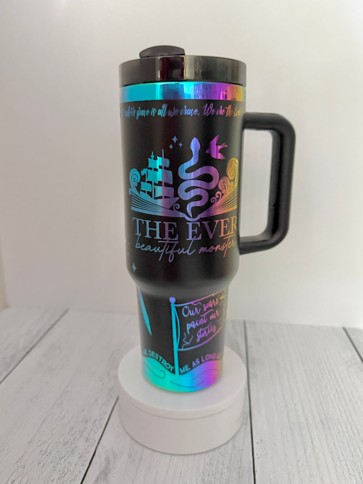 A black insulated Ever King themed travel mug with a handle, featuring holographic rainbow artwork and text including 'THE EVER: beautiful monster' and quotes from the book by LJ Andrews, shown on a white pedestal against a light gray background.
