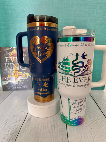 Two Ever King themed handled insulated tumblers displayed on a white pedestal against a teal backdrop and light wood surface. The left tumbler is dark blue with gold lettering reading 'Hello Love' and small gold decorative accents. The right is white with rainbow engraved details 'The Ever: beautiful monster'