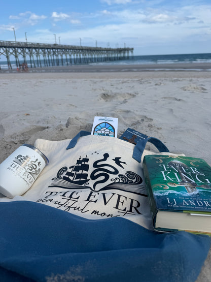 Tote bag with The Ever design and 'The Ever King' book by LJ Andrews on a beach with a pier in the background