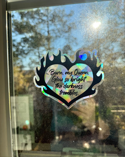 Flaming heart suncatcher decal on a window with the quote "Burn, my Queen. Glow so bright the darkness trembles."
