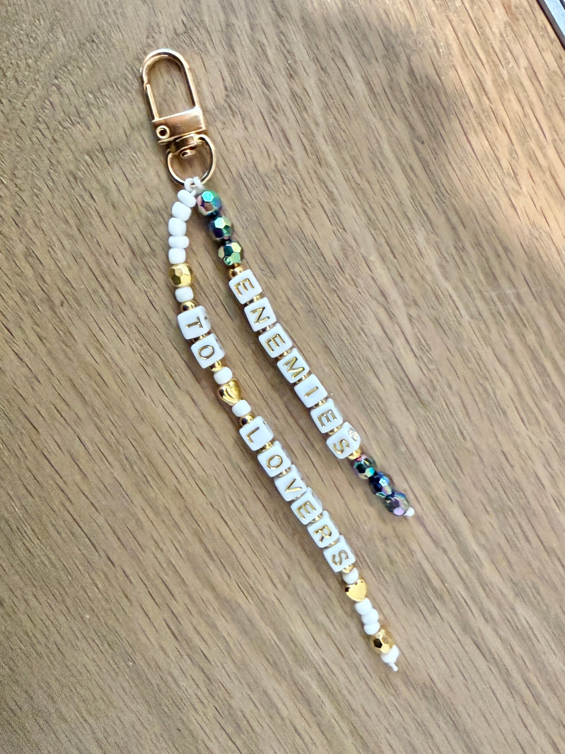 A beaded keychain or bag charm with a gold-tone lobster clasp, featuring white letter beads spelling 'ENEMIES TO LOVERS' alongside gold and iridescent beads, displayed on a light wood surface.
