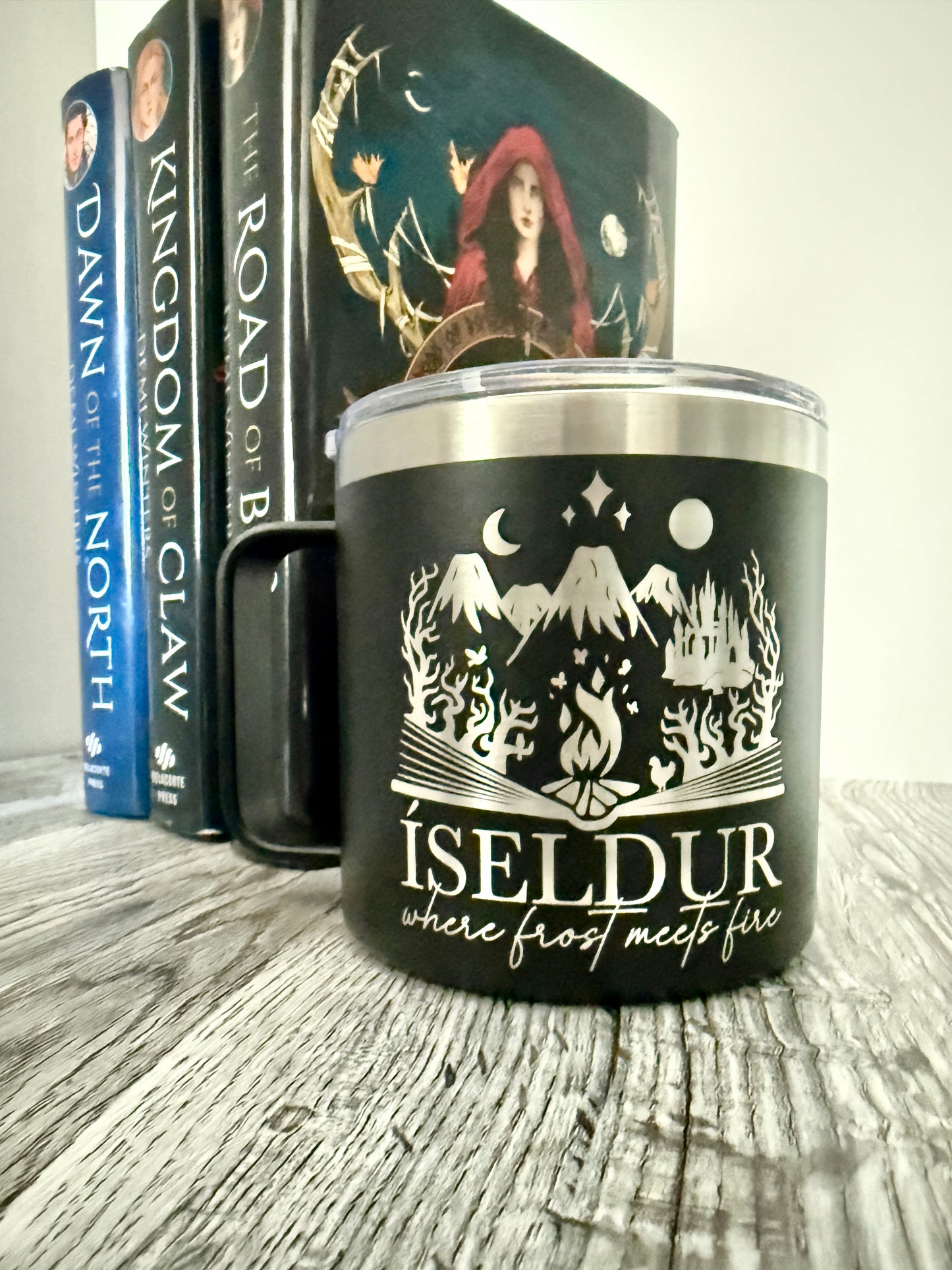 Black camp mug with 'Iseldur: where frost meets fire' bookish fantasy design, placed on a wooden surface with Demi Winters' Ashen series books in the background.