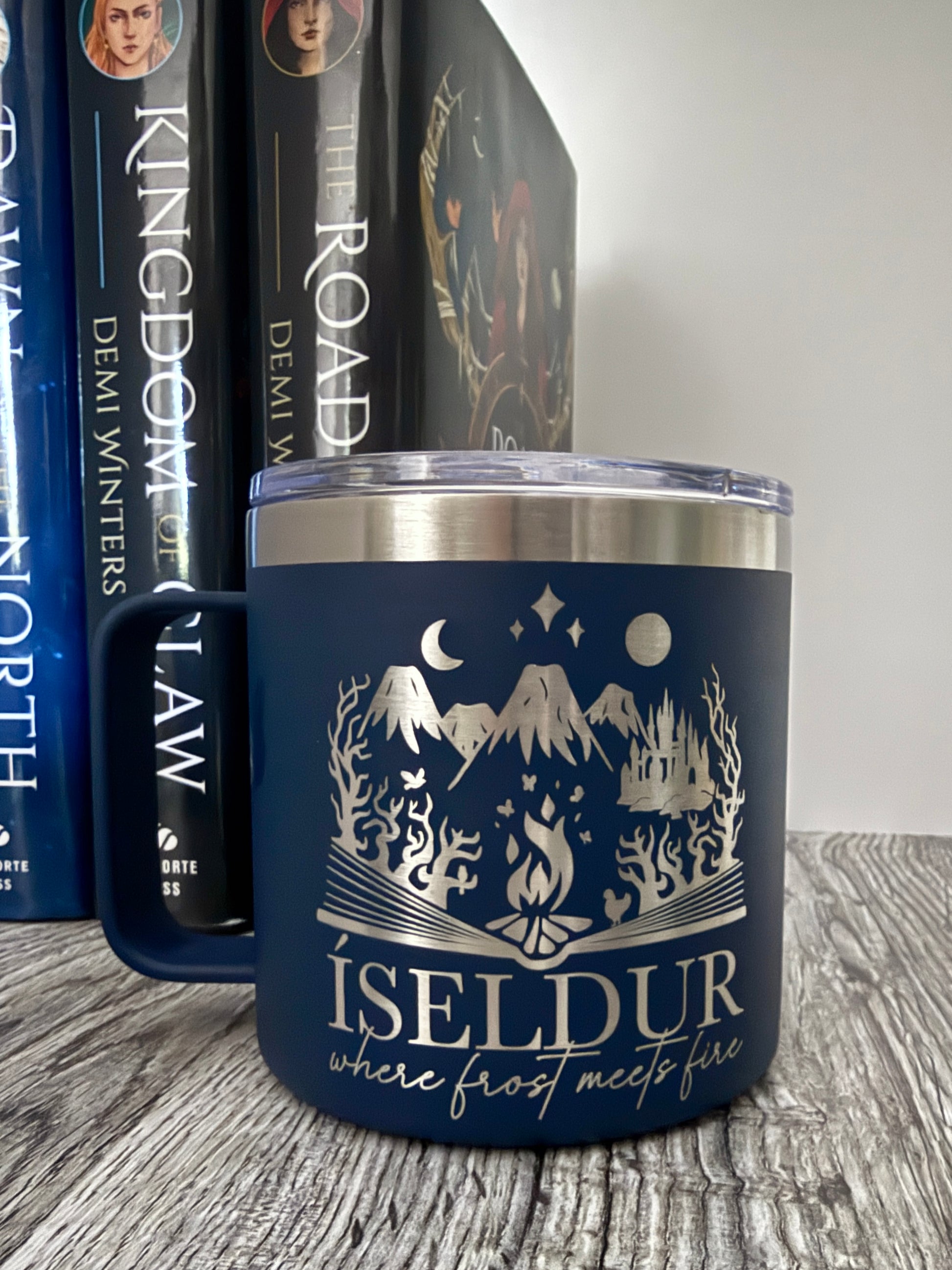 Navy camp mug with 'Iseldur: where frost meets fire' bookish design, placed on a rustic wood surface with Demi Winters' Road of Bones books in the background.