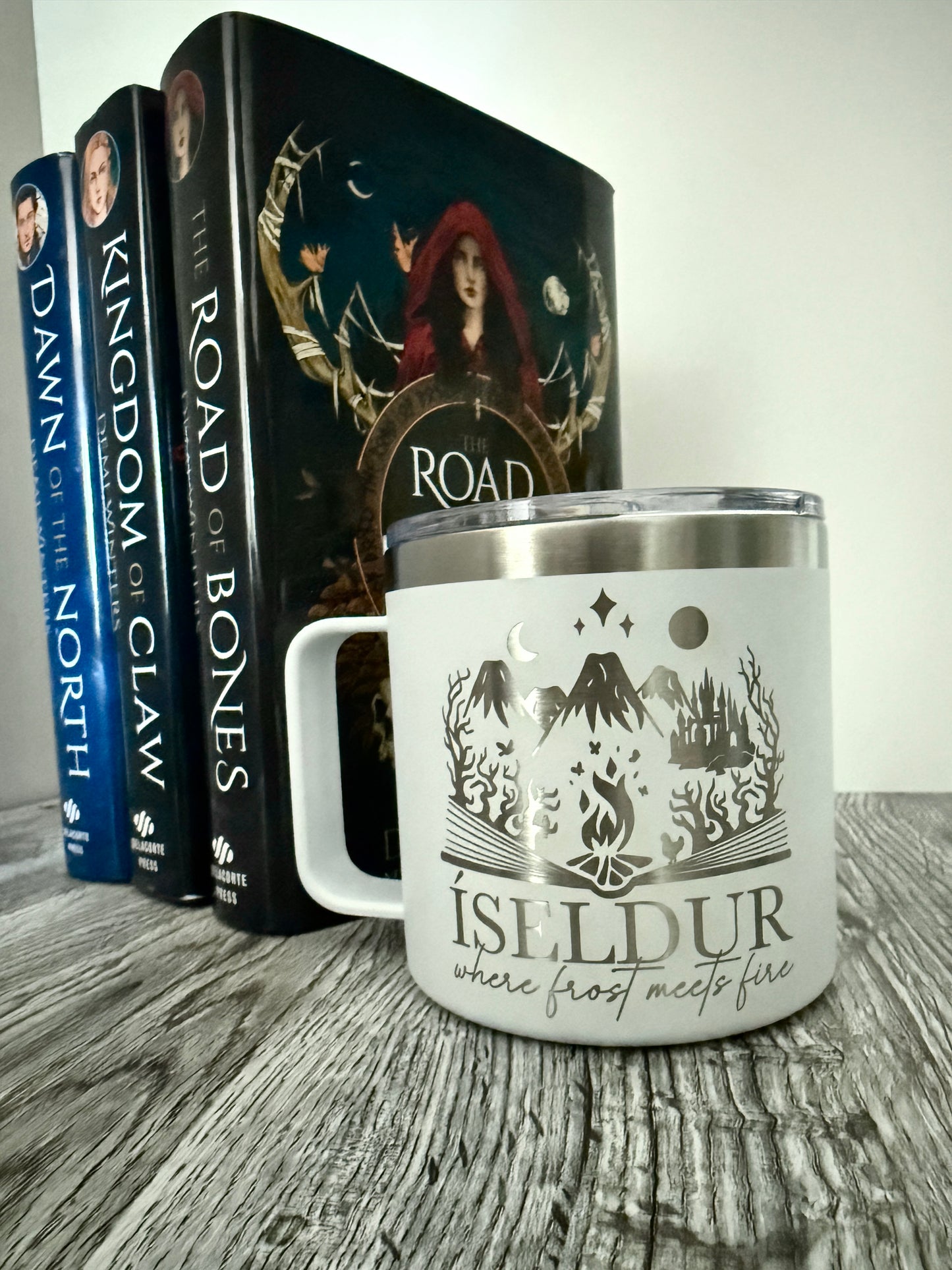 White camp mug with 'Isildur: where frost meets fire' bookish design next to Demi Winters' Road of Bones books on a wooden surface