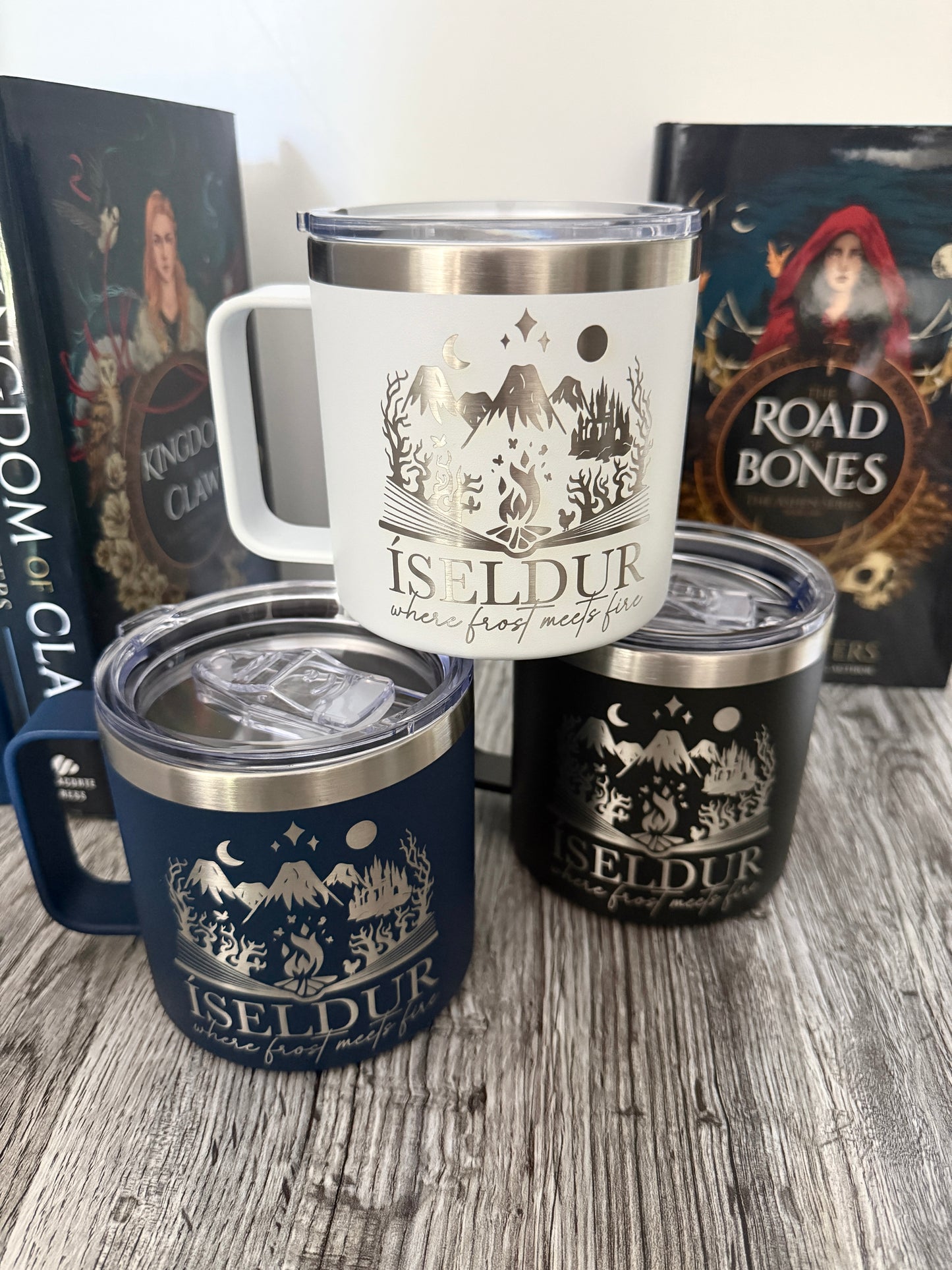 Three camp mugs with 'Iseldur: where frost meets fire' bookish design on a wooden surface with Demi Winters' Road of Bones and Kingdom of Claw books in the background.