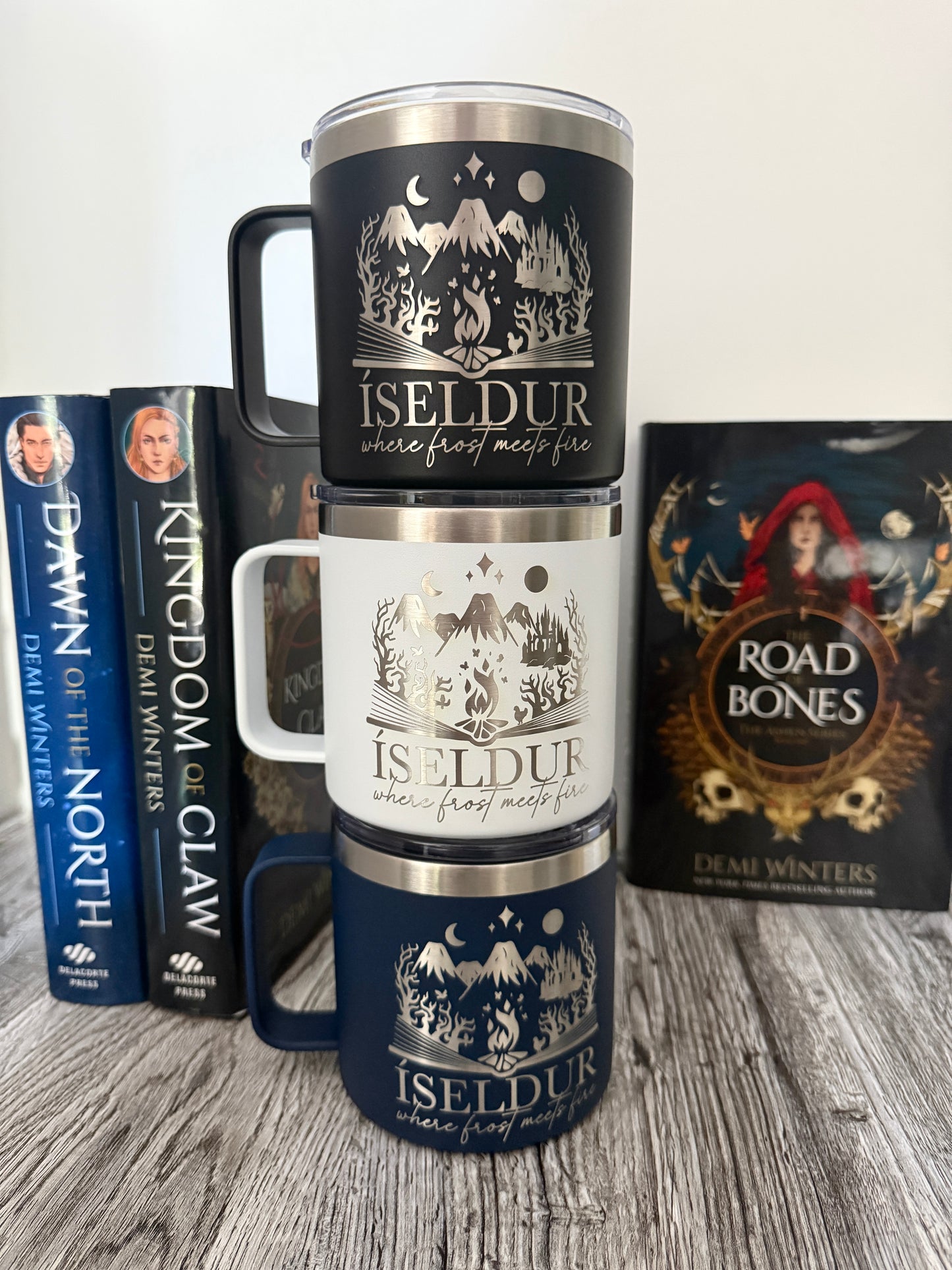 Three stacked camp mugs with 'Iseldur: where frost meets fire' bookish design on a wooden surface with Demi Winters' Ashen series books in the background.