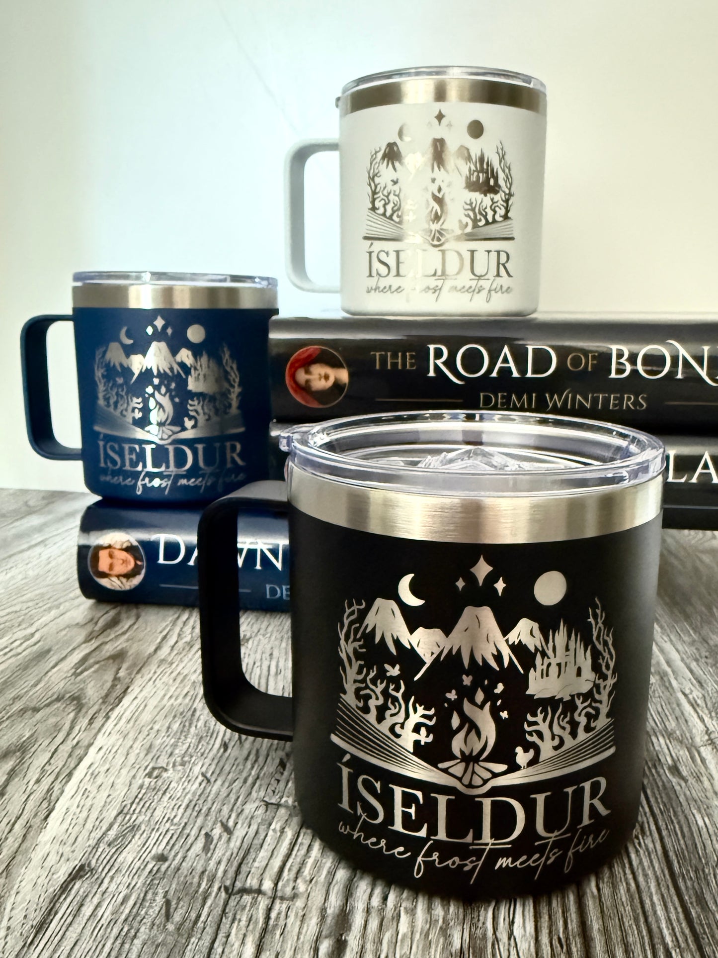 Three insulated camp mugs with 'Iseldur: where frost meets fire' bookish design on a wooden surface with Demi Winters' The Road of Bones book in the background.