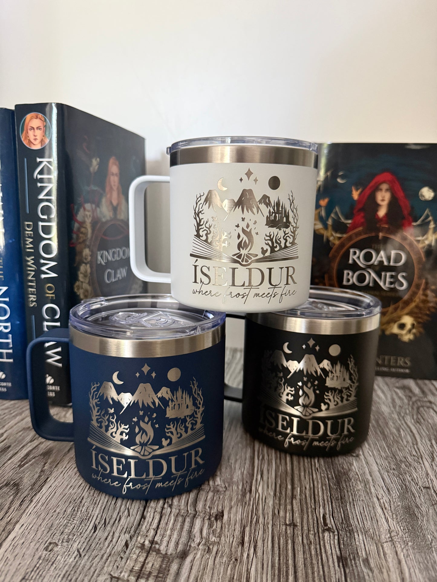 Three insulated travel mugs with 'Iseldur: where frost meets fire' bookish design on a wooden surface, with Demi Winters' The Road of Bones and Kingdom of Claw books in the background.