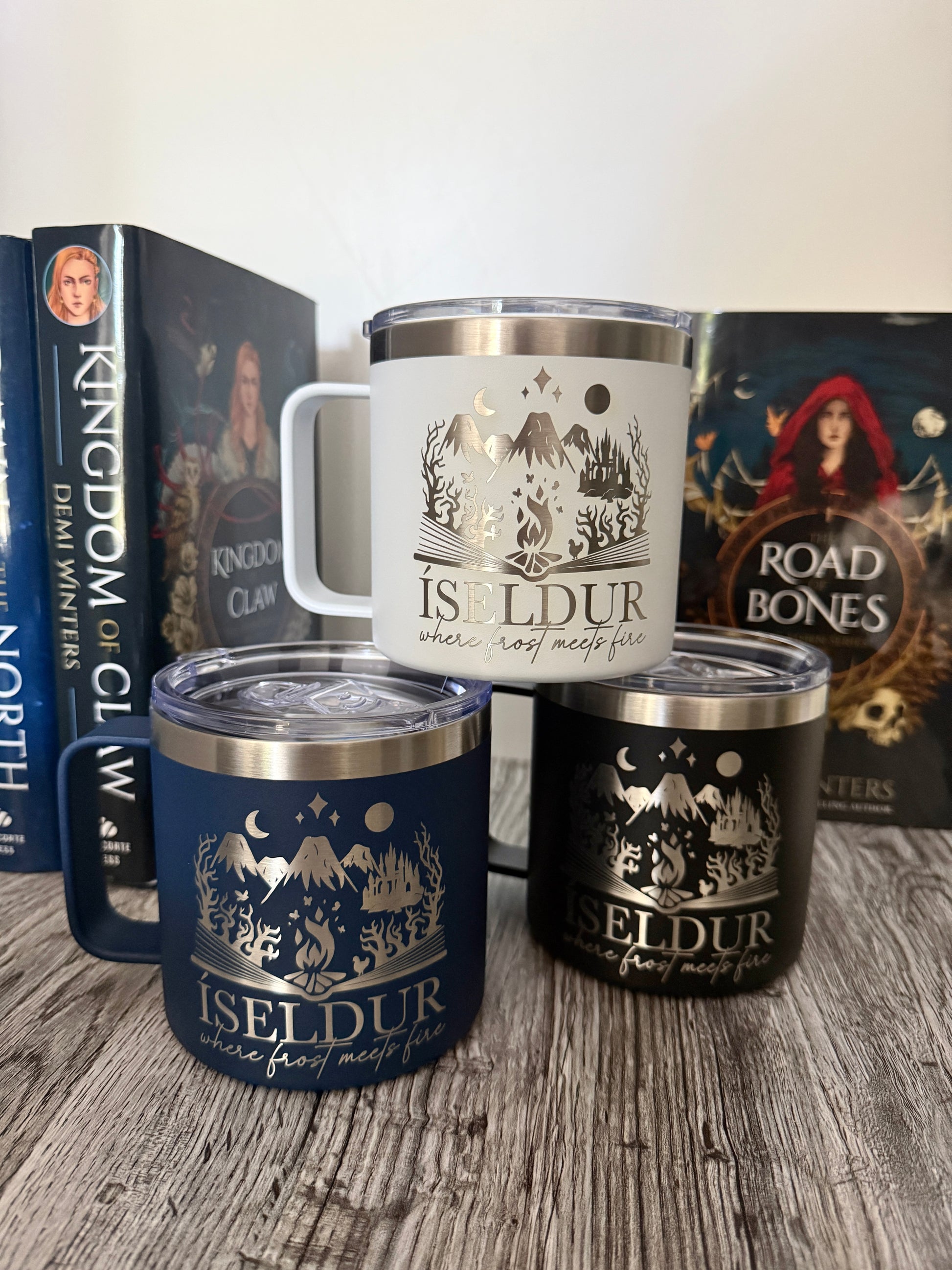 Three insulated travel mugs with 'Iseldur: where frost meets fire' bookish design on a wooden surface, with Demi Winters' The Road of Bones and Kingdom of Claw books in the background.