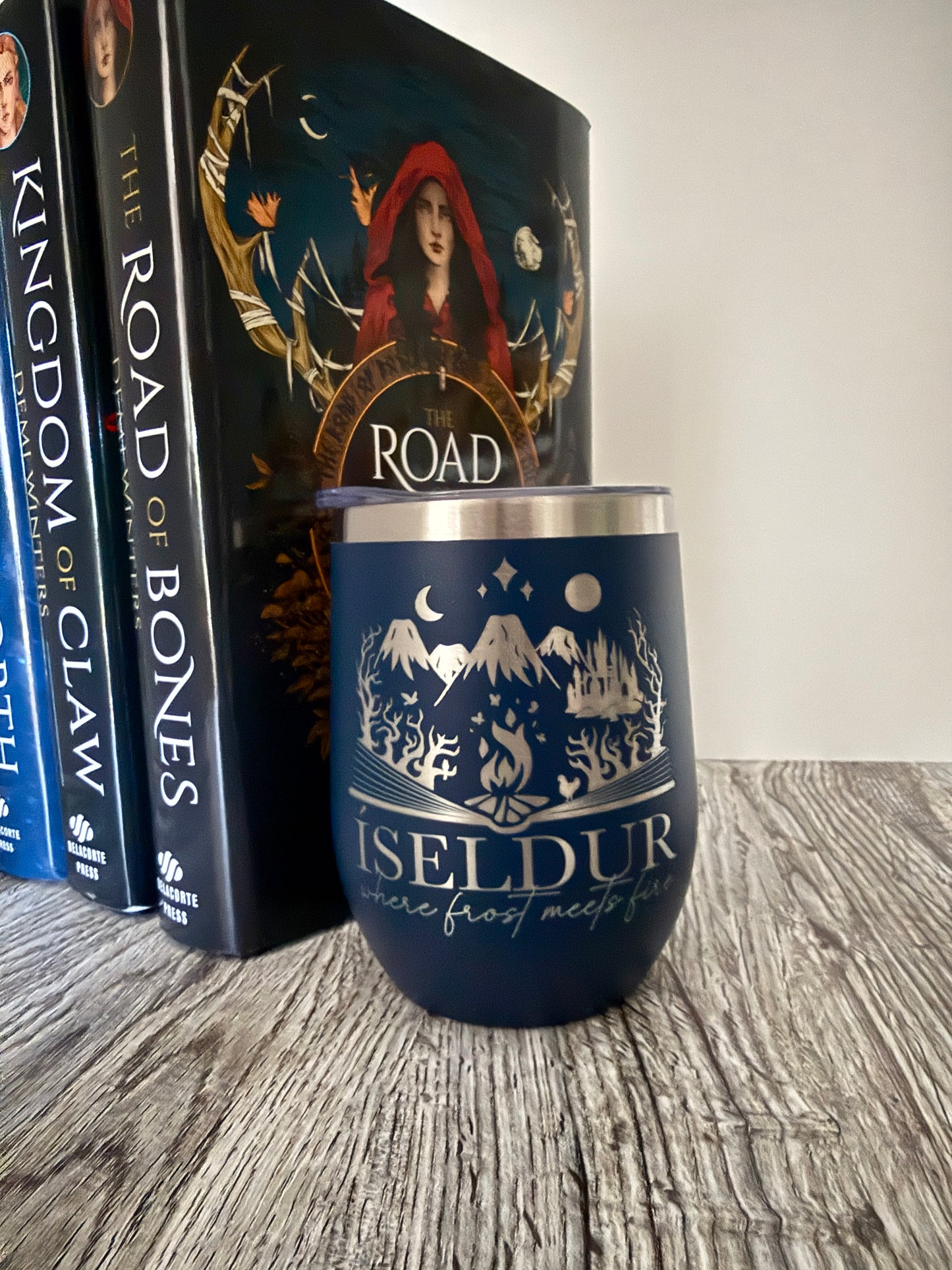 Navy blue wine tumbler with mountain design and 'Iseldur: where frost meets fire' text on a wooden surface with Demi Winters' The Road of Bones books in the background.