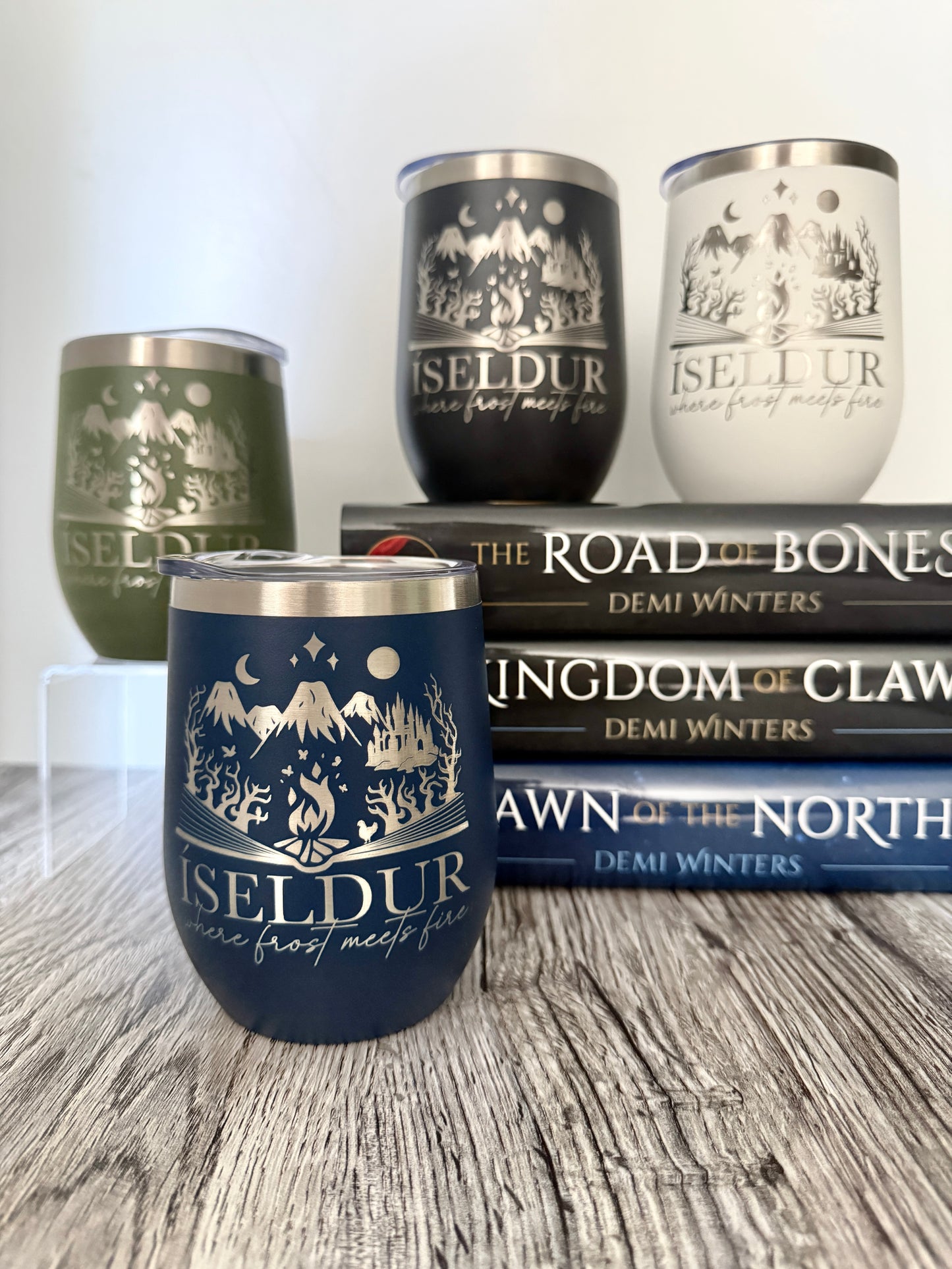 Four insulated tumblers with 'Iseldur: where frost meets fire' fantasy design on a wooden surface with Demi Winters' Ashen series books in the background.