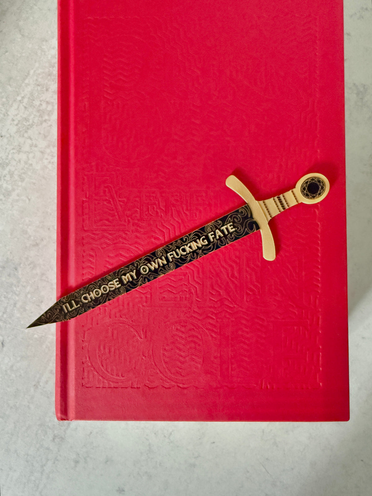 A decorative sword bookmark with a black and gold patterned blade and a gold hilt, placed diagonally across a red hardcover book. The blade features a quote about choosing your own fate.