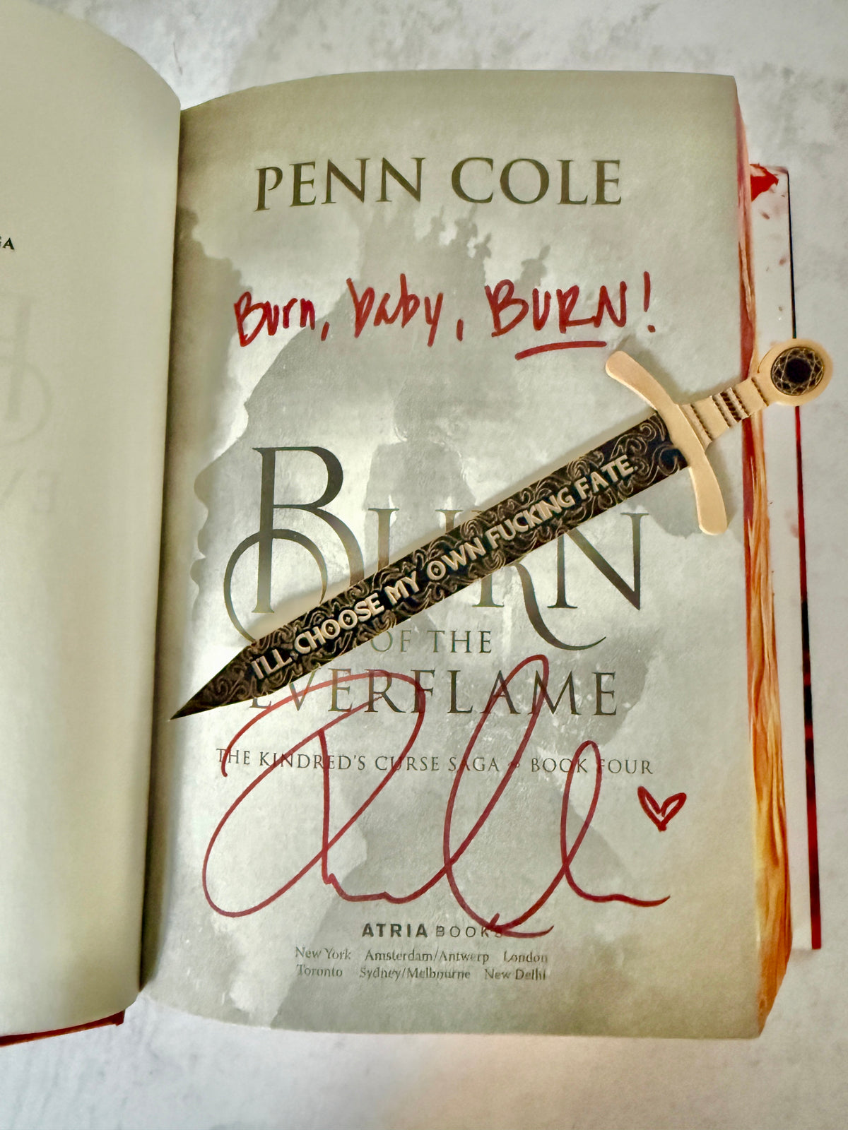An open copy of the book 'Burn of the Everflame' by Penn Cole, signed by the author, showing the title page with a decorative sword bookmark that features a quote about choosing your own fate.