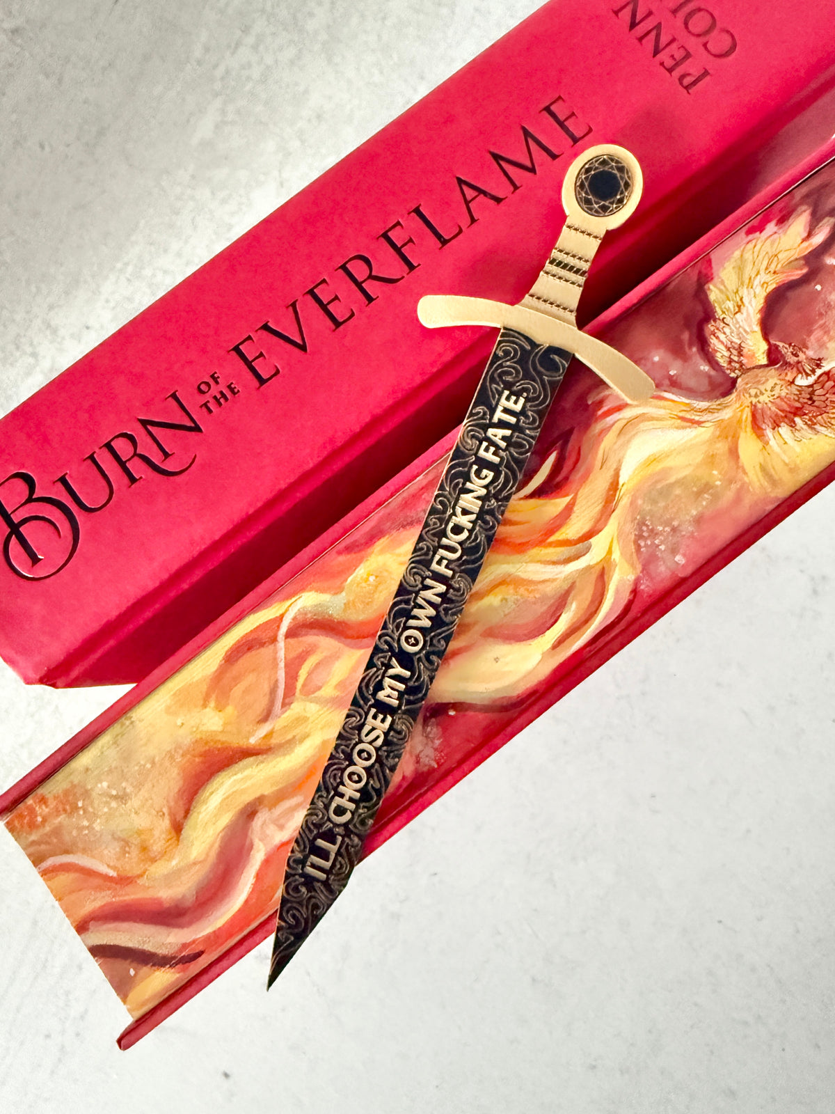 A decorative sword bookmark with a black engraved blade with gold hilt, engraved with a quote about choosing your own fate, displayed diagonally over a red book 'BURN OF THE EVERFLAME' and a fiery phoenix design painted edges.