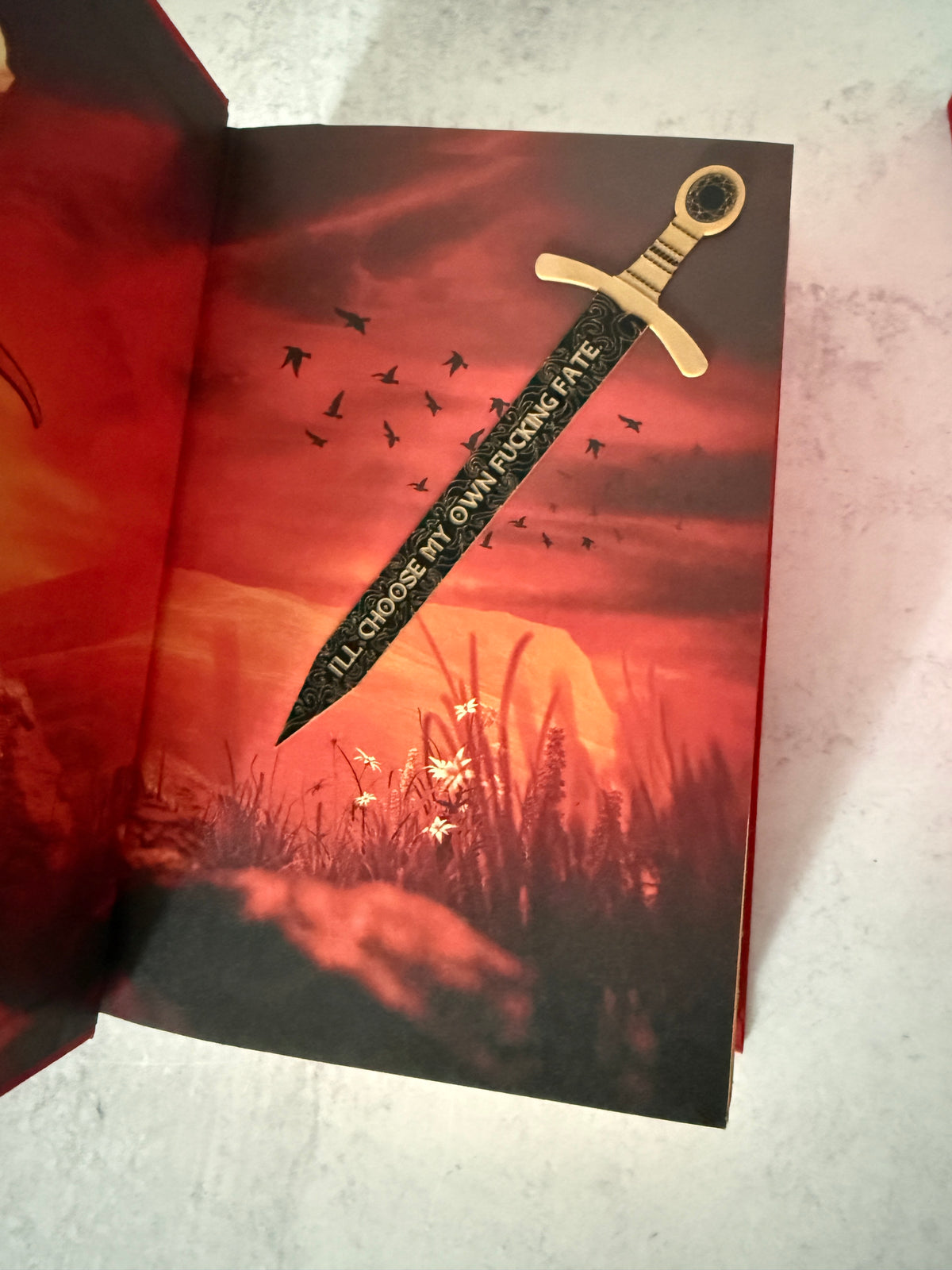 A decorative sword bookmark with a gold-colored hilt and a black blade printed with the quote about choosing your own fate, shown inserted into an open book of Burn of the Everflame with a red and black fantasy illustration.
