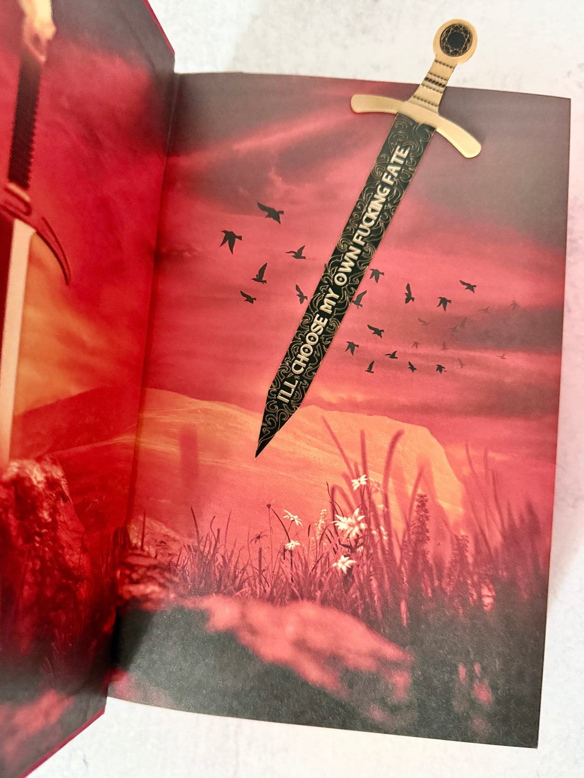 A decorative sword bookmark with a gold-colored hilt and a black blade printed with the quote about choosing your own fate, shown inserted into an open book of Burn of the Everflame with a red and black fantasy illustration.