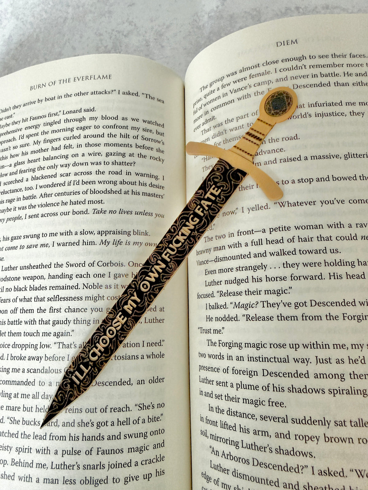 Black and gold sword bookmark engraved with a quote about choosing your own fate, resting on an open book of 'Burn of the Everflame.'