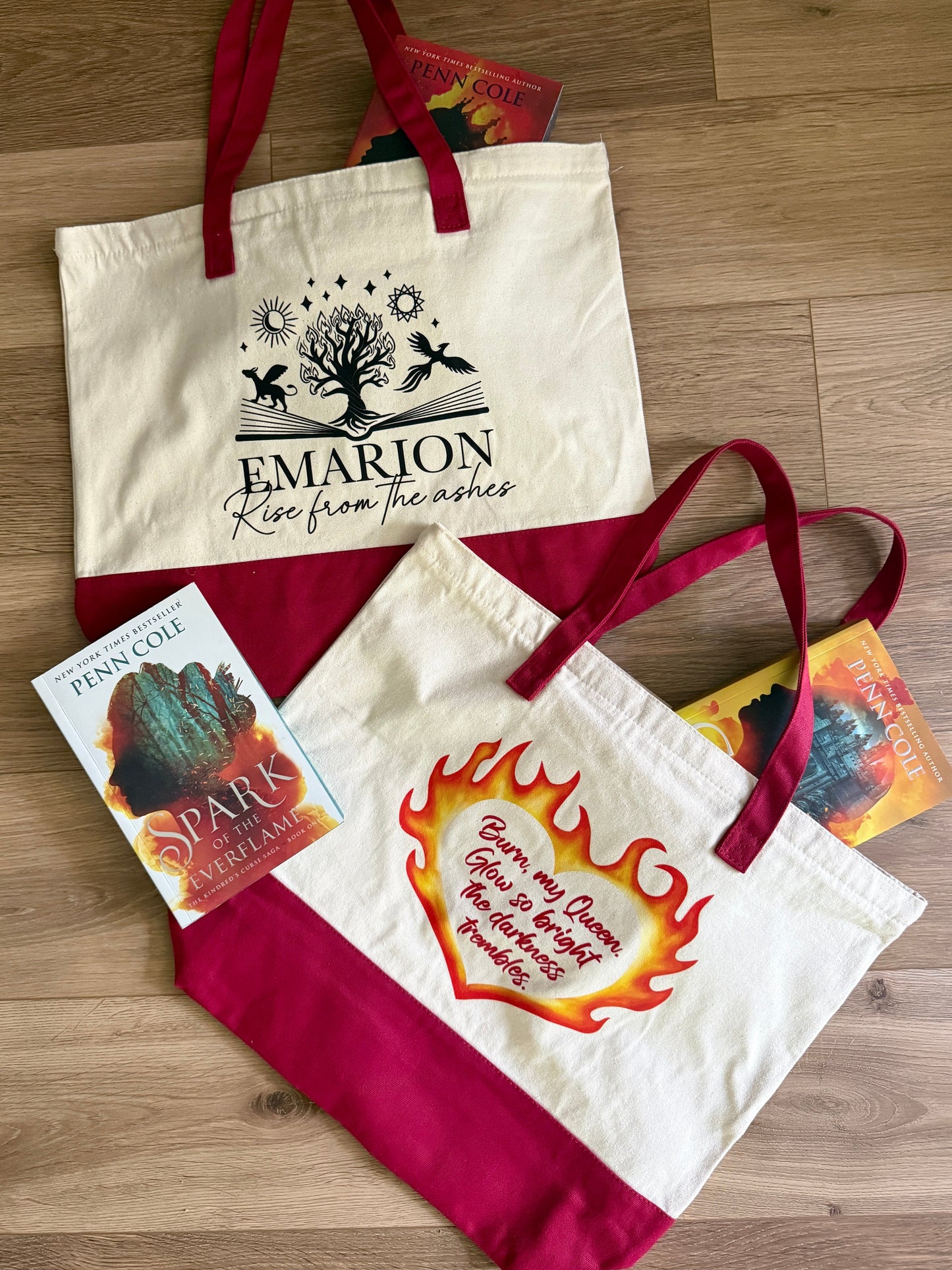 Two Kindred's Curse Saga tote bags with printed designs inspired by Penn Cole's books on a wooden surface. Bags feature quotes "Burn my Queen. Glow so bright the darkness trembles." and "rise from the ashes."