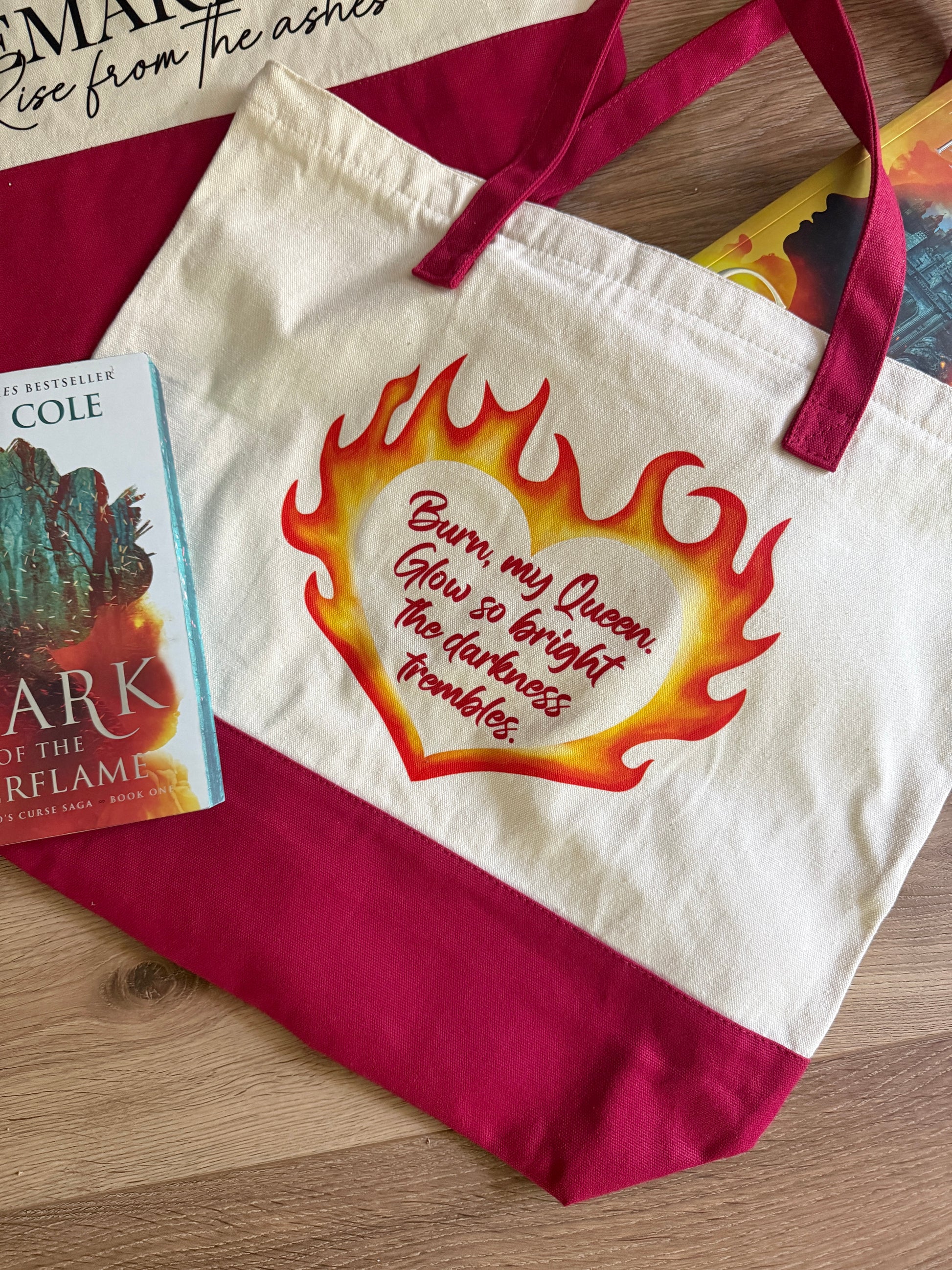Tote bag with flaming heart design and quote "Burn my Queen. Glow so bright the darkness trembles" inspired by Penn Cole's Kindred's Curse Saga next to a Spark of the Everflame book on a wooden surface