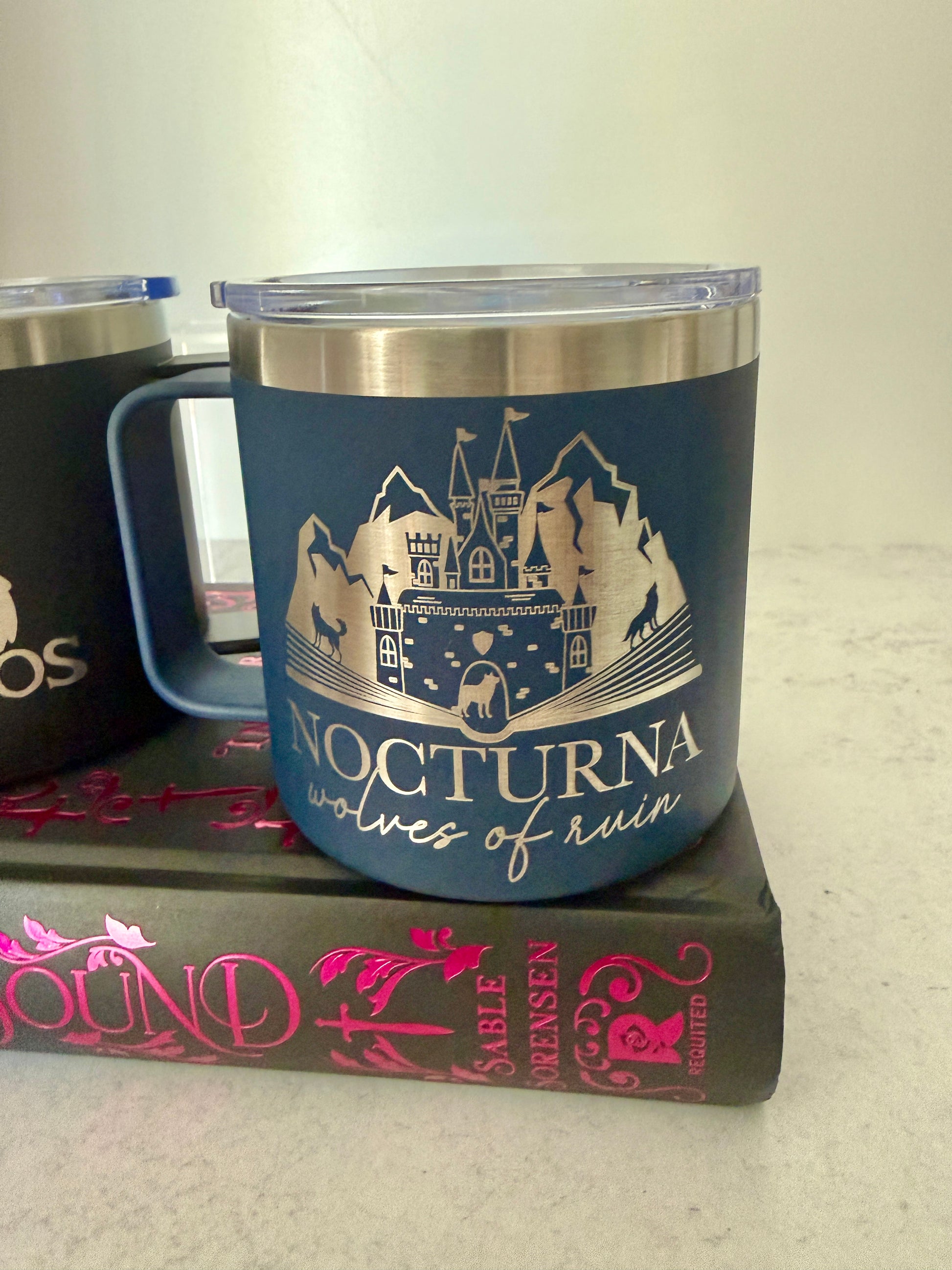 Blue tumbler with 'Nocturna' wolves of ruin castle and mountain design on a Dire Bound book by Sable Sorensen