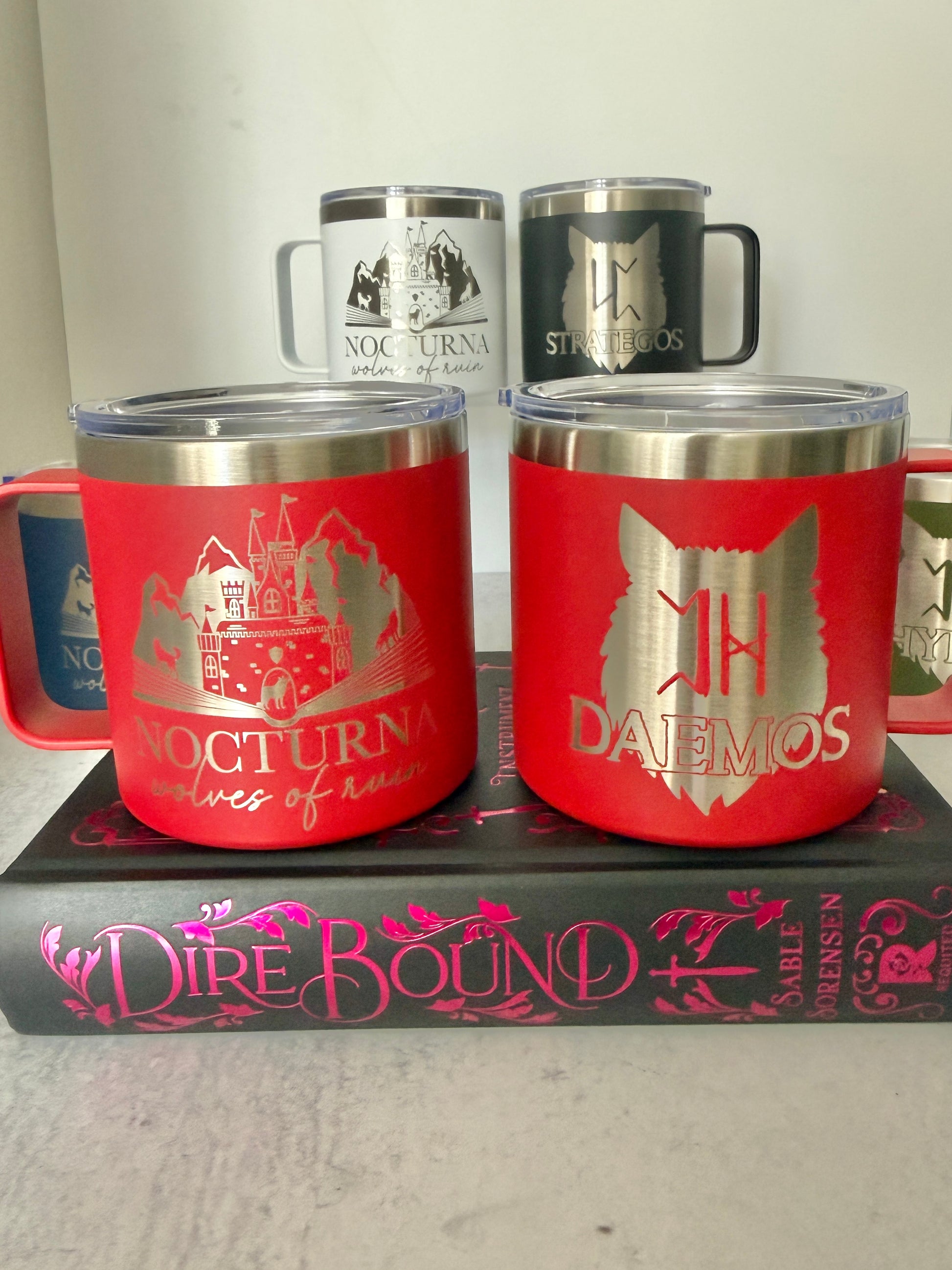 Set of insulated mugs with Nocturna castle and wolf designs on a book titled 'Dire Bound'.