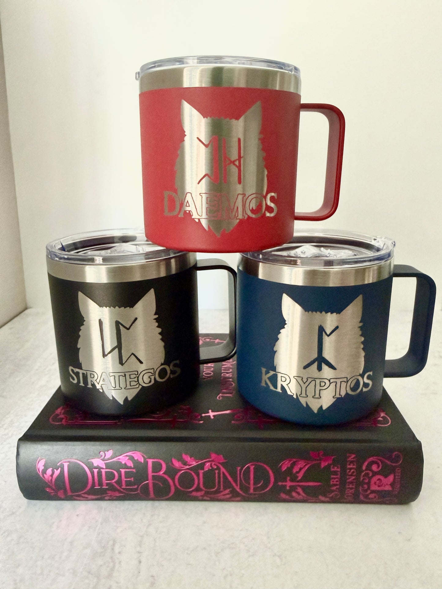 Three insulated mugs with wolf pack crests on a book titled 'Dire Bound'.