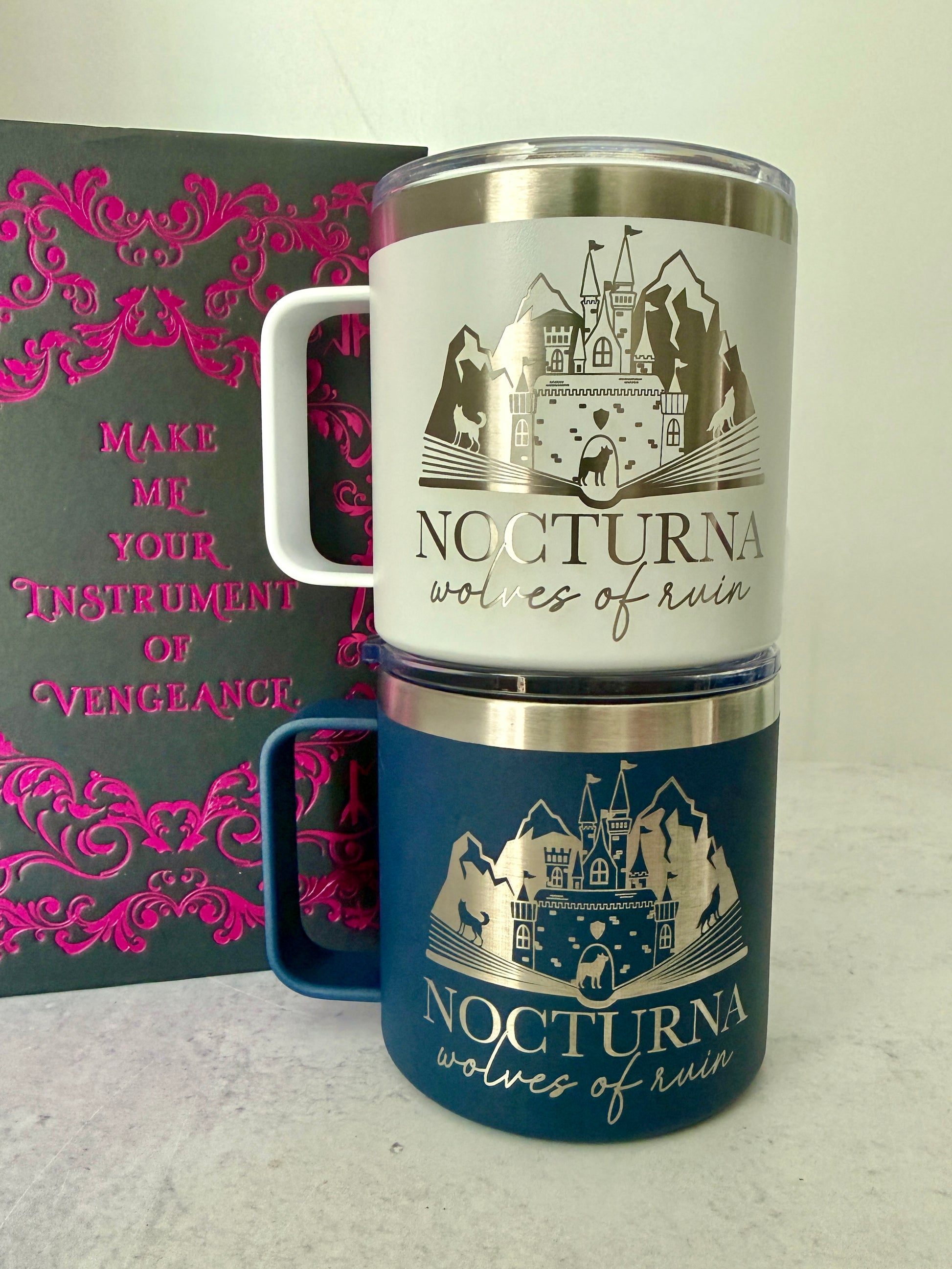 Two insulated camp mugs with 'Nocturna wolves of ruin' design on a light gray background with a book that has the quote "make me your instrument of vengeance."
