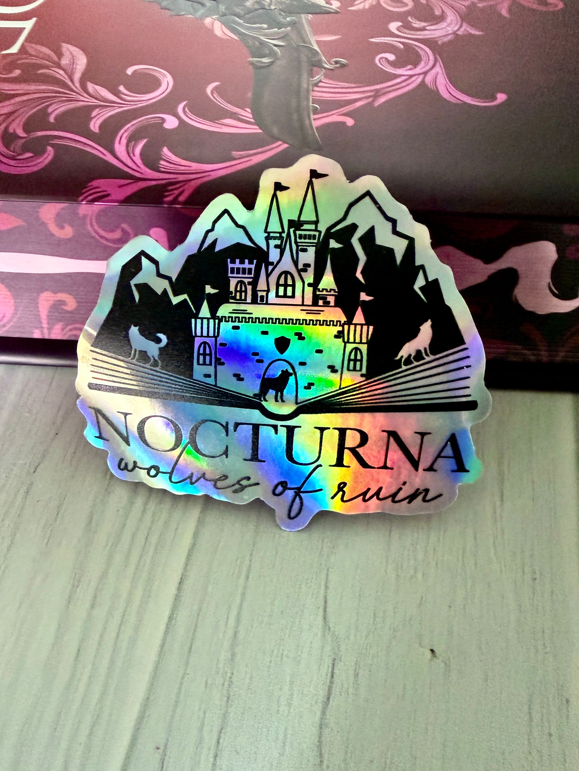 Holographic sticker with castle design and 'Nocturna Wolves of Ruin' text on a wooden surface with Dire Bound book.