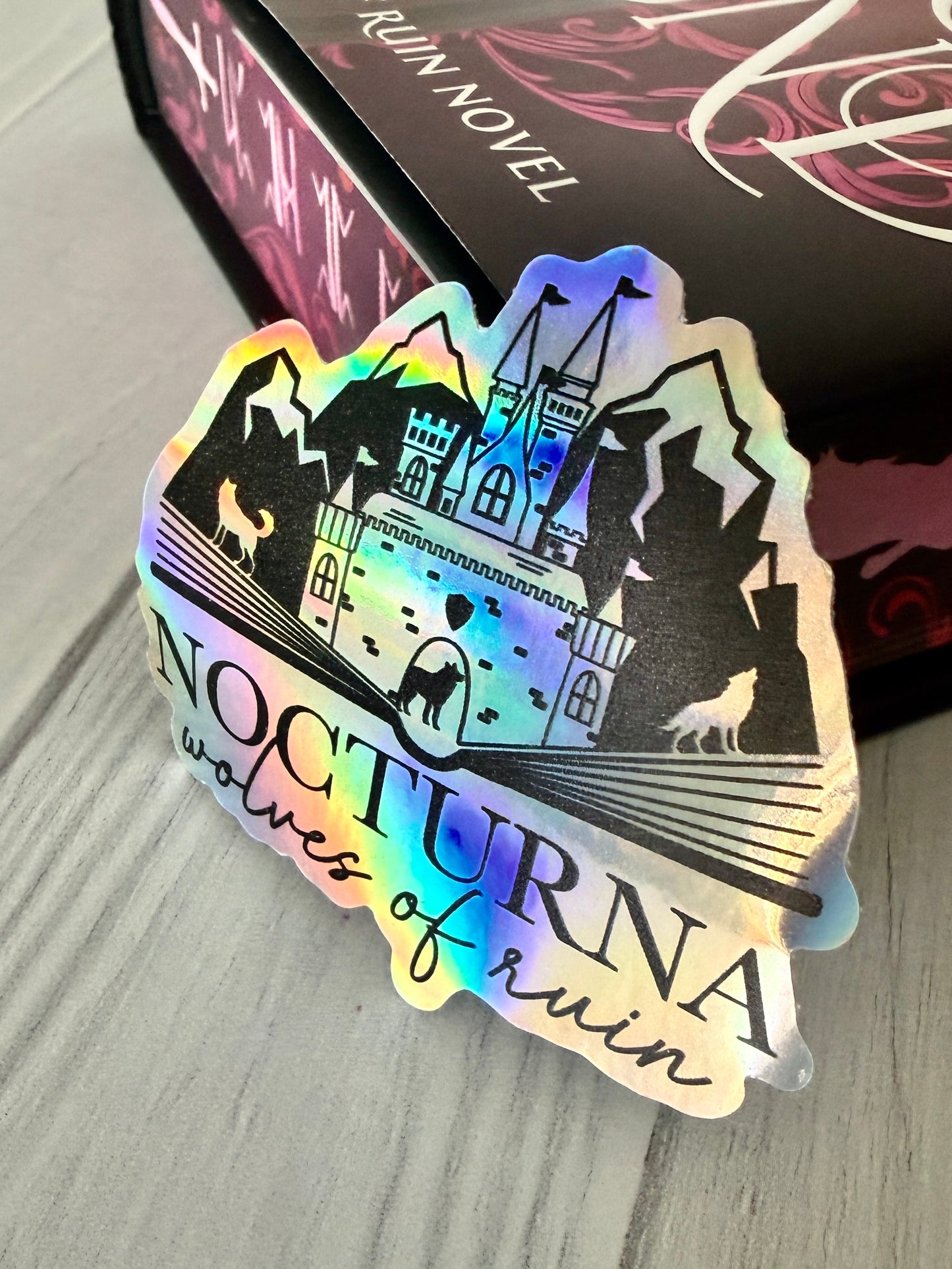 Holographic sticker with castle design and 'Nocturna Wolves of Ruin' text on a wooden surface, leaning on a copy of Dire Bound.
