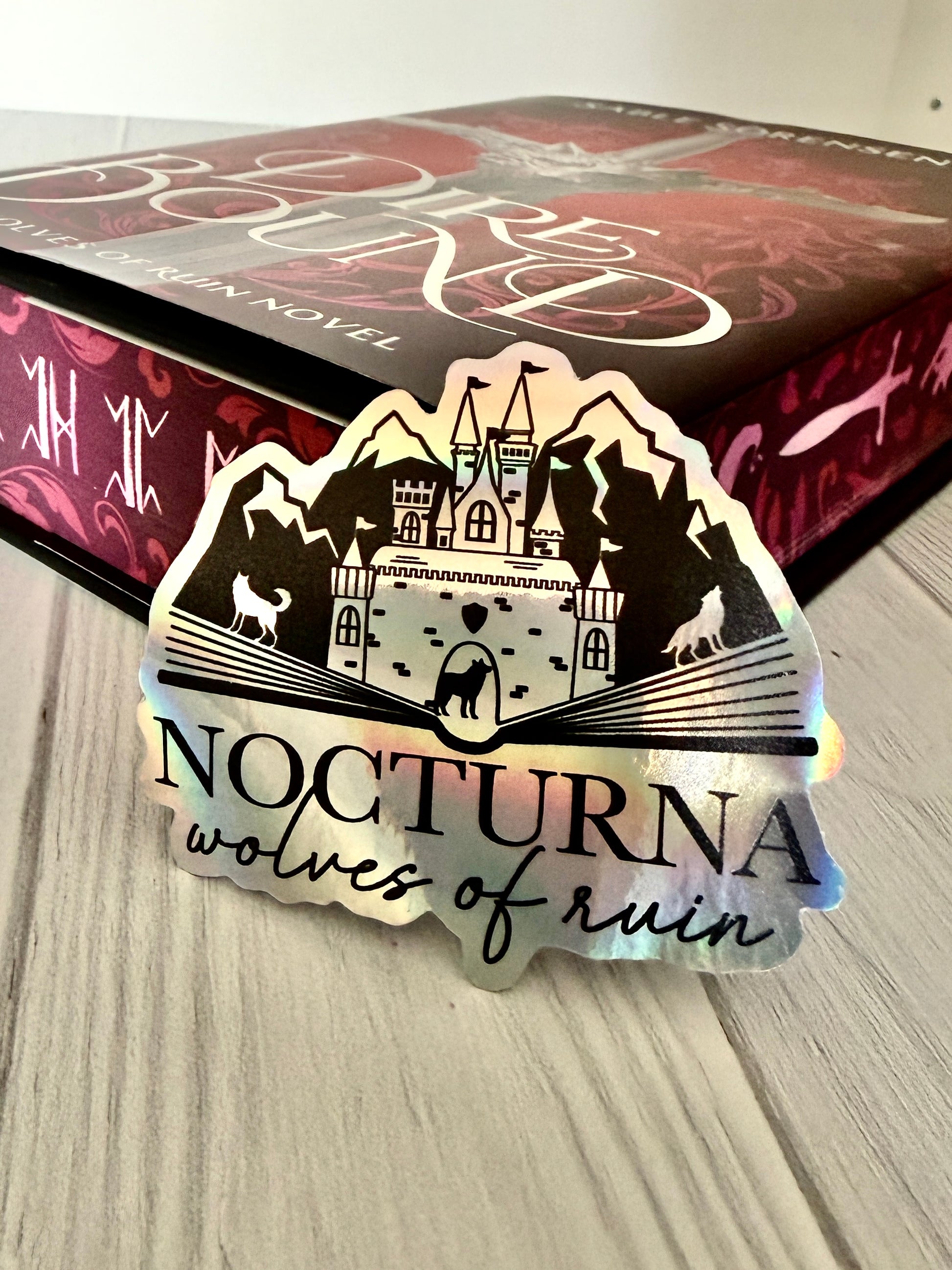 Sticker with 'Nocturna wolves of ruin' design on a textured wood surface with Dire Bound book.