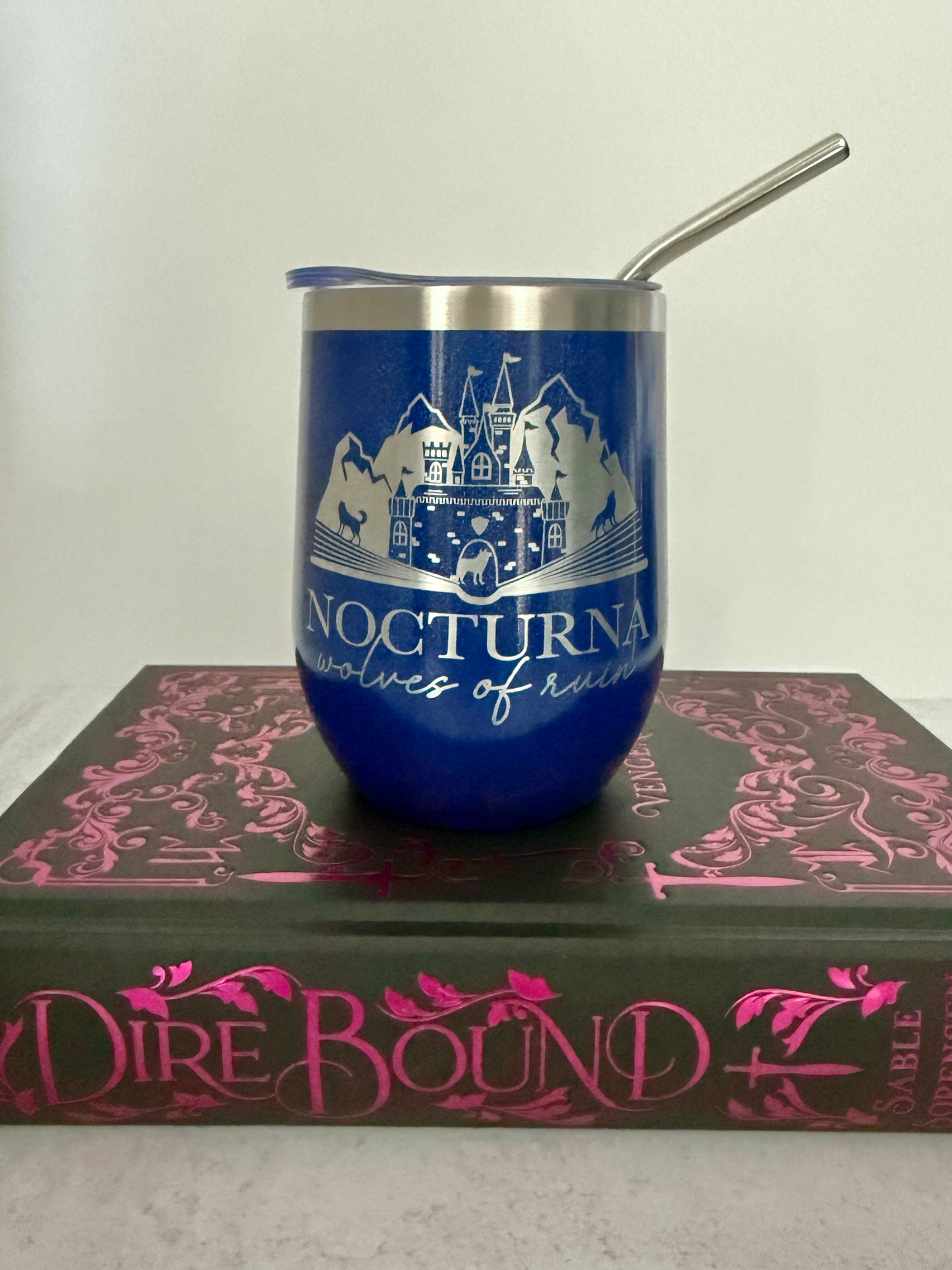 Blue tumbler with a castle design and "Nocturna: wolves of ruin" on top of a book titled 'Dire Bound'.
