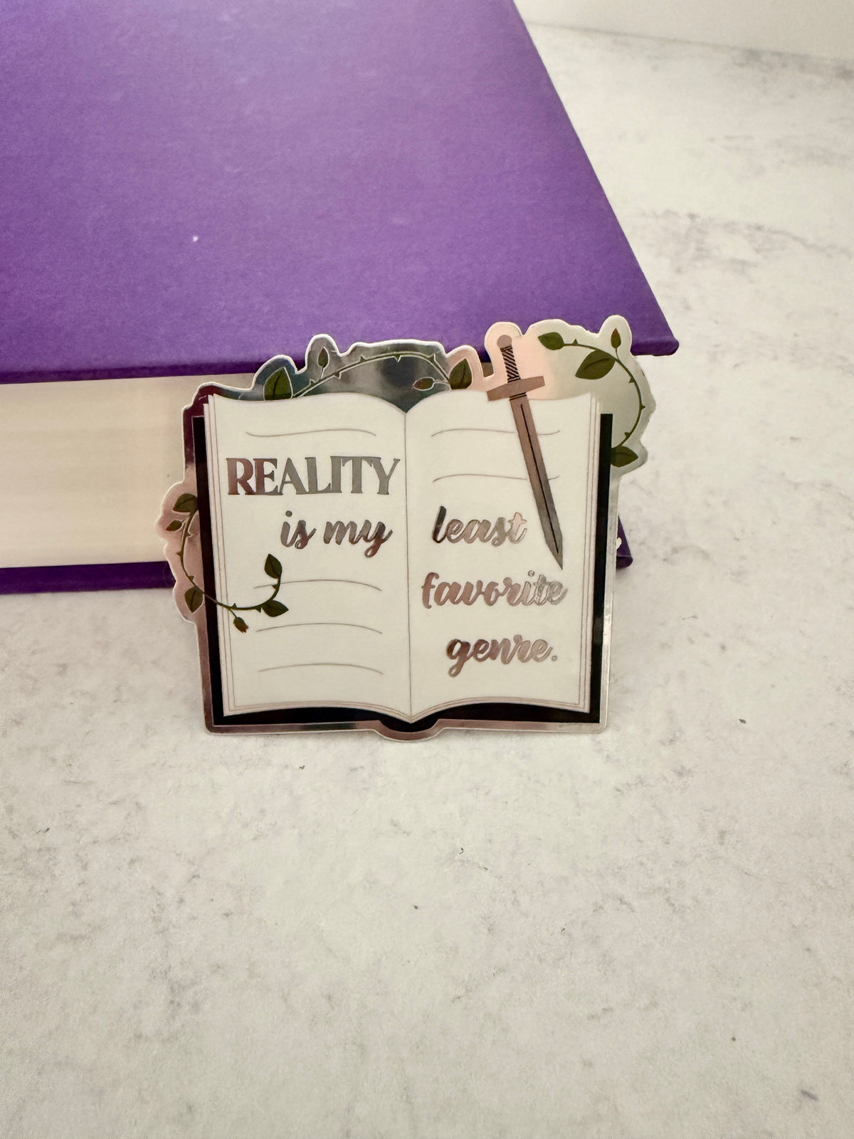 Open book sticker with metallic lettering 'reality is my least favorite genre,' a sword, and thorned vines, leaning against a purple book