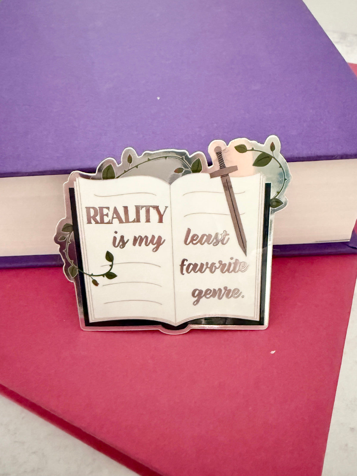 Open book sticker with metallic lettering 'reality is my least favorite genre,' a sword, and thorned vines, tilted on purple and red books.