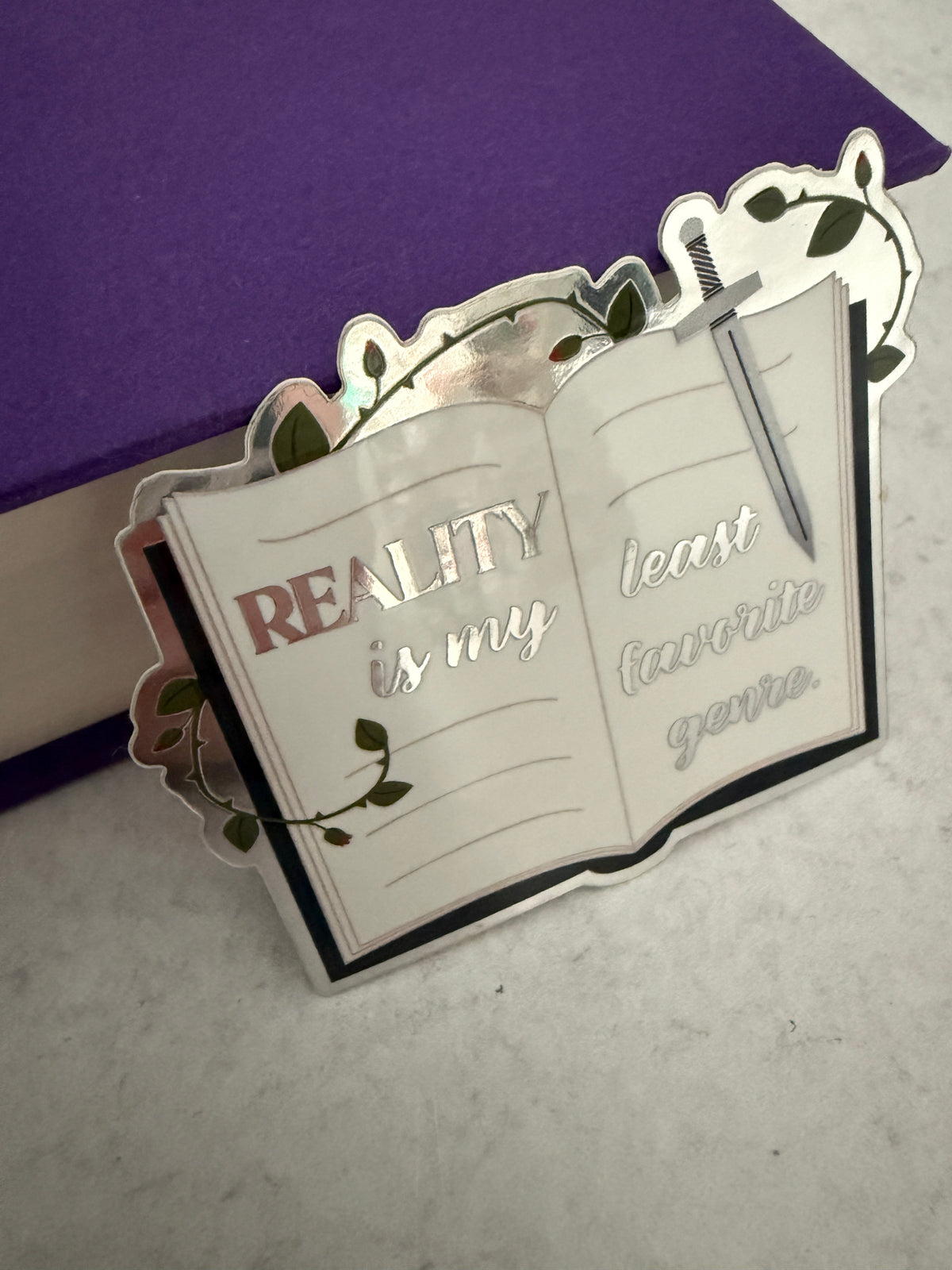 Open book sticker with metallic lettering 'reality is my least favorite genre,' a sword, and thorned vines, leaning against a purple book