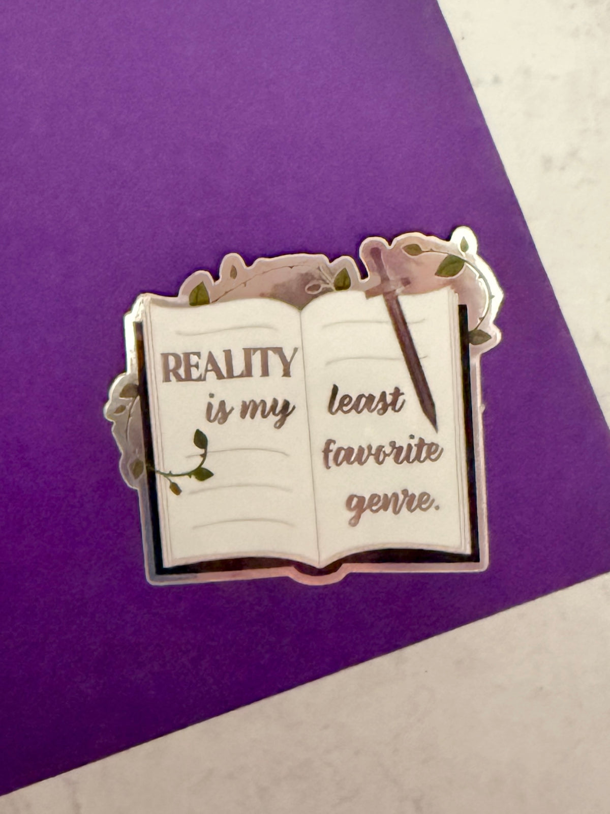 Open book sticker with metallic lettering 'reality is my least favorite genre,' a sword, and thorned vines, laying on top of a purple book
