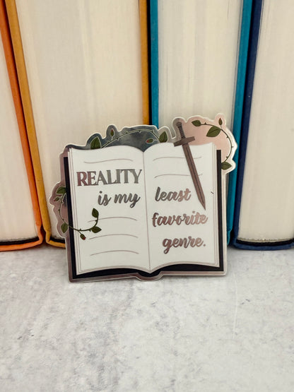 Reality is my least favorite genre metallic book sticker leaning against hardcover books with orange, teal, and navy spines