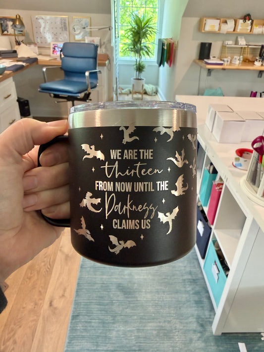 Person holding a black mug with engraved text "we are the thirteen from now until the darkness claims us" with wyverns flying around it, shown in an office setting