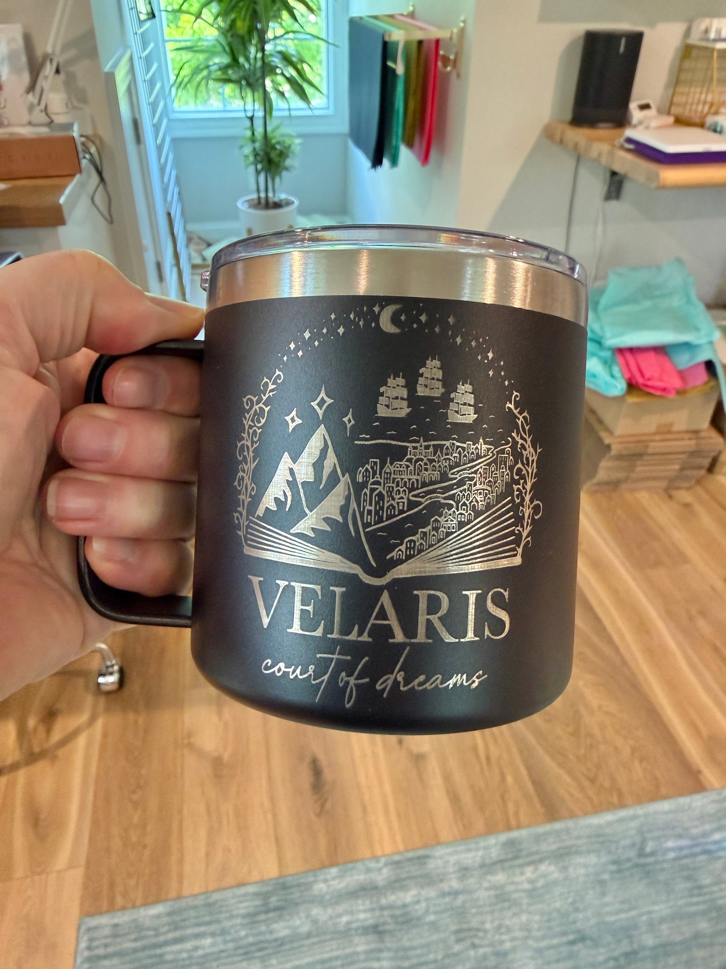 Black mug with 'Velaris: court of dreams' engraved on it and held by a hand in a studio setting