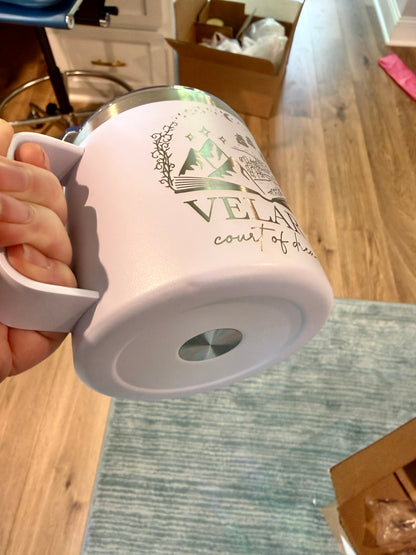 Person holding a white mug with 'Velaris: court of dreams' engraving showing the bottom of the mug