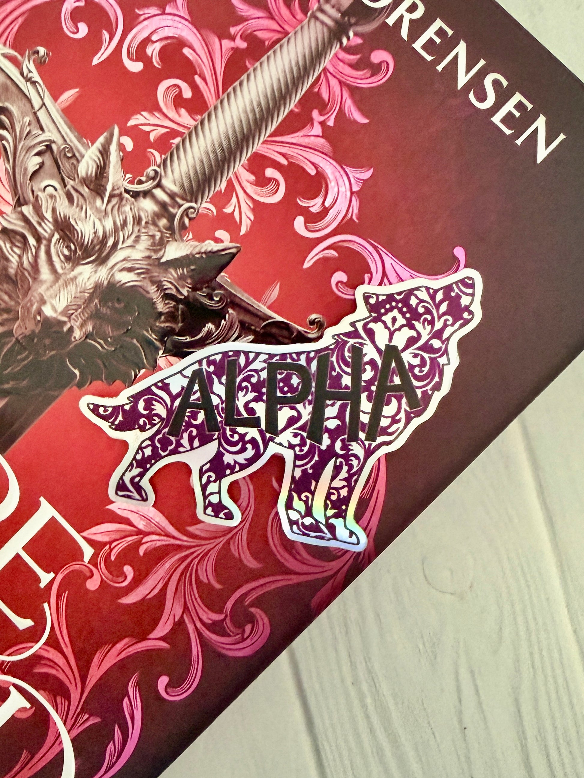 Wolf shaped holographic sticker with 'Alpha' text on a copy of Dire Bound by Sable Sorensen