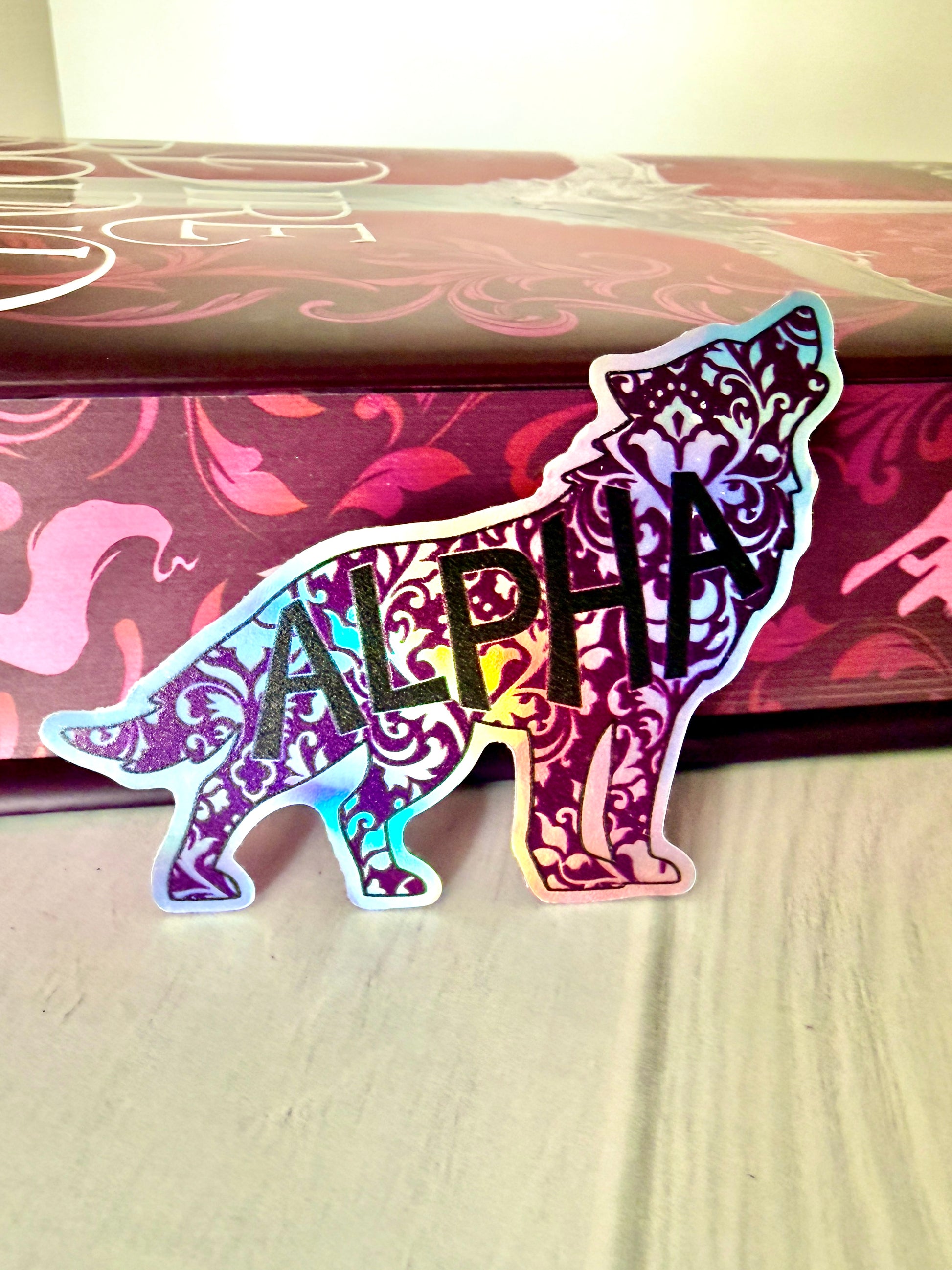 Decorative wolf shaped die cut holographic sticker with 'ALPHA' leaning against the sprayed edges of Dire Bound.