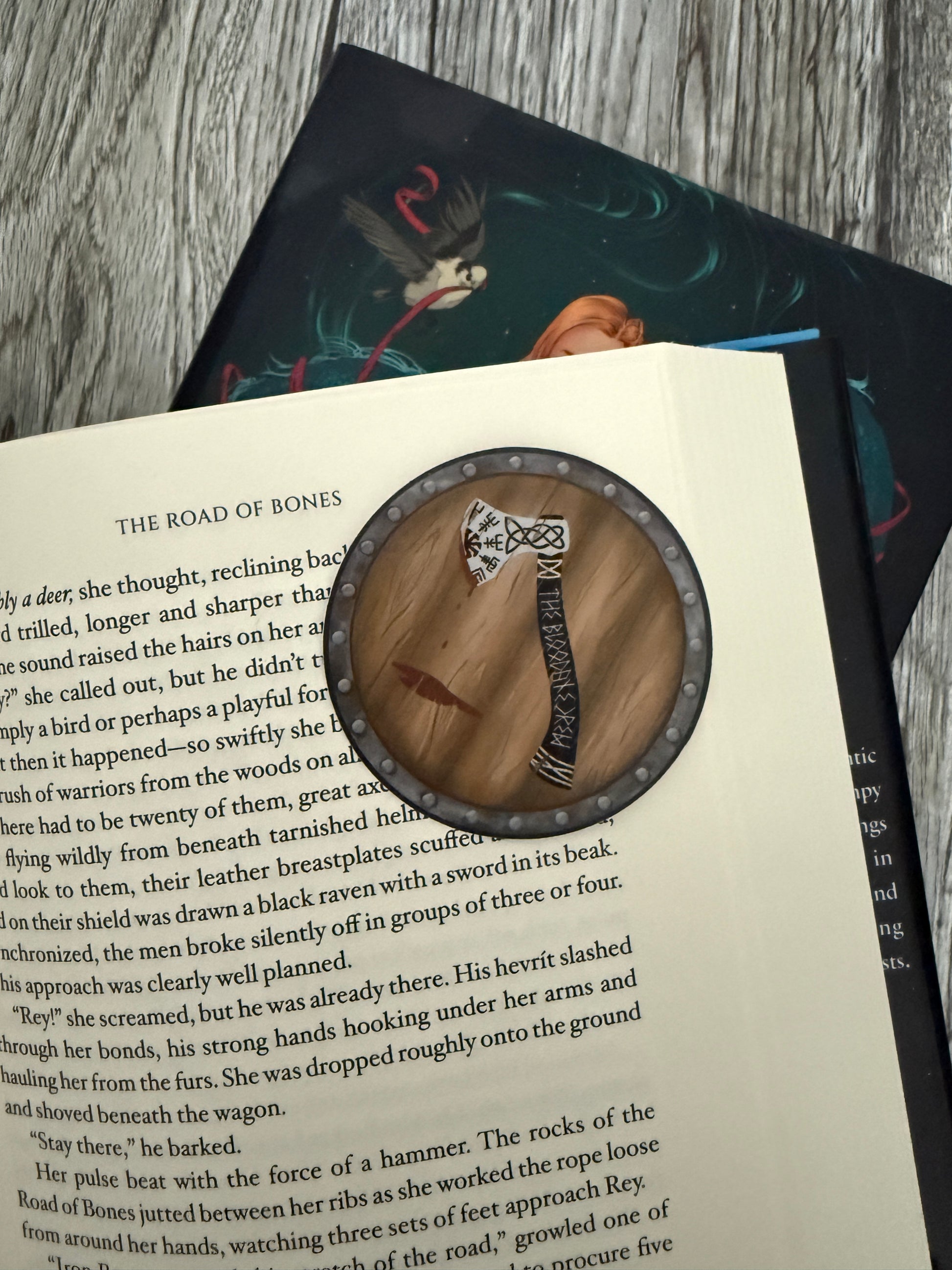 Round sticker hand drawn with a viking axe on a shield, laying on an open book of The Road of Bones by Demi Winters