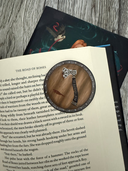 Round sticker hand drawn with a viking axe on a shield, laying on an open book of The Road of Bones by Demi Winters
