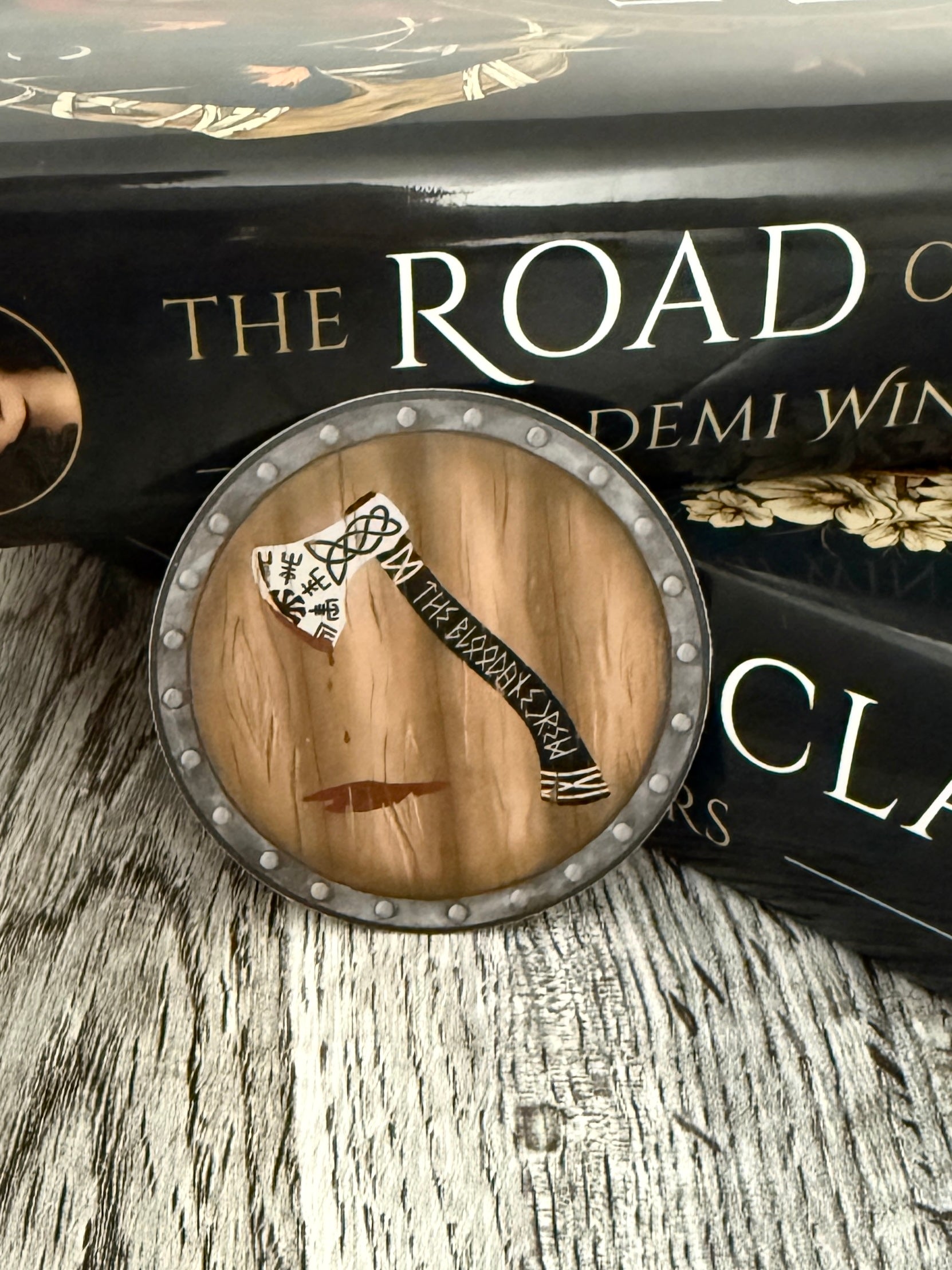 Die cut sticker in the shape of a shield with an viking axe dripping with blood painted on it and The Bloodaxe Crew written in runes down the handle, leaning against the Road of Bones book by Demi Winters