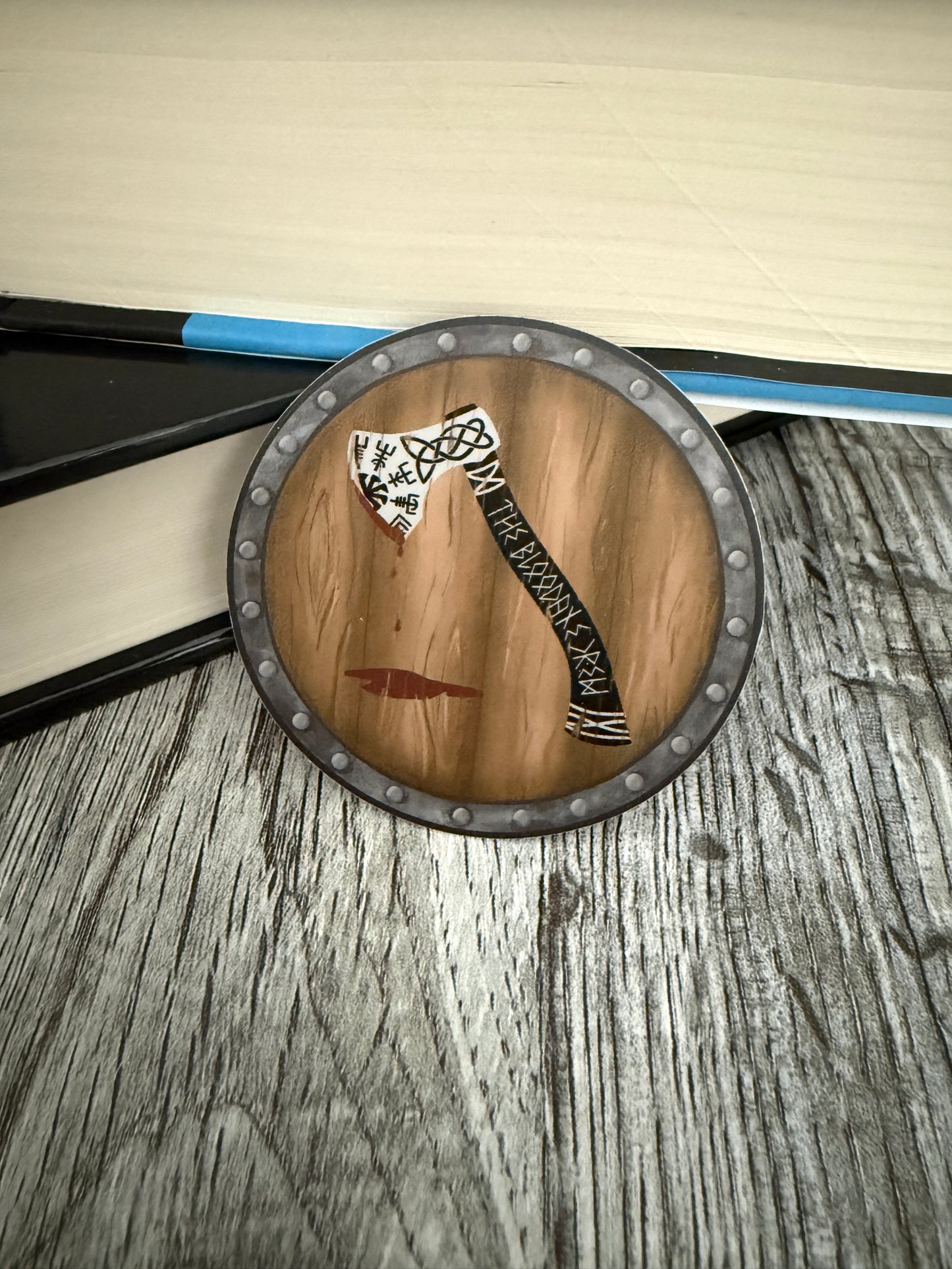 Round viking shield sticker featuring a decorative axe with The Bloodaxe Crew in runes down the handle, leaning against books on a wooden background