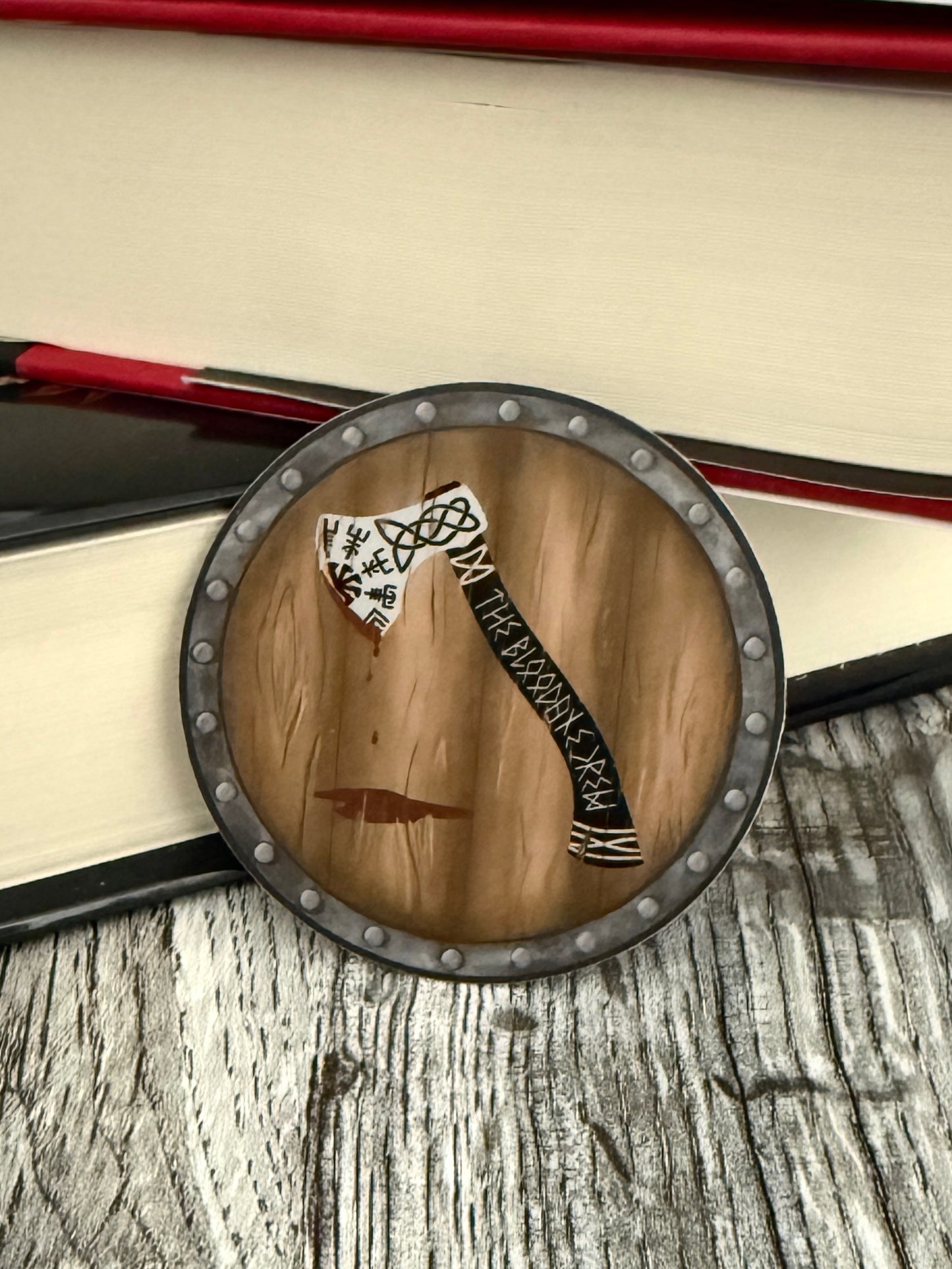 Round viking shield die cut sticker featuring an axe that reads The Bloodaxe Crew in runes down the handle, leaning against books, inspired by The Road of Bones by Demi Winters