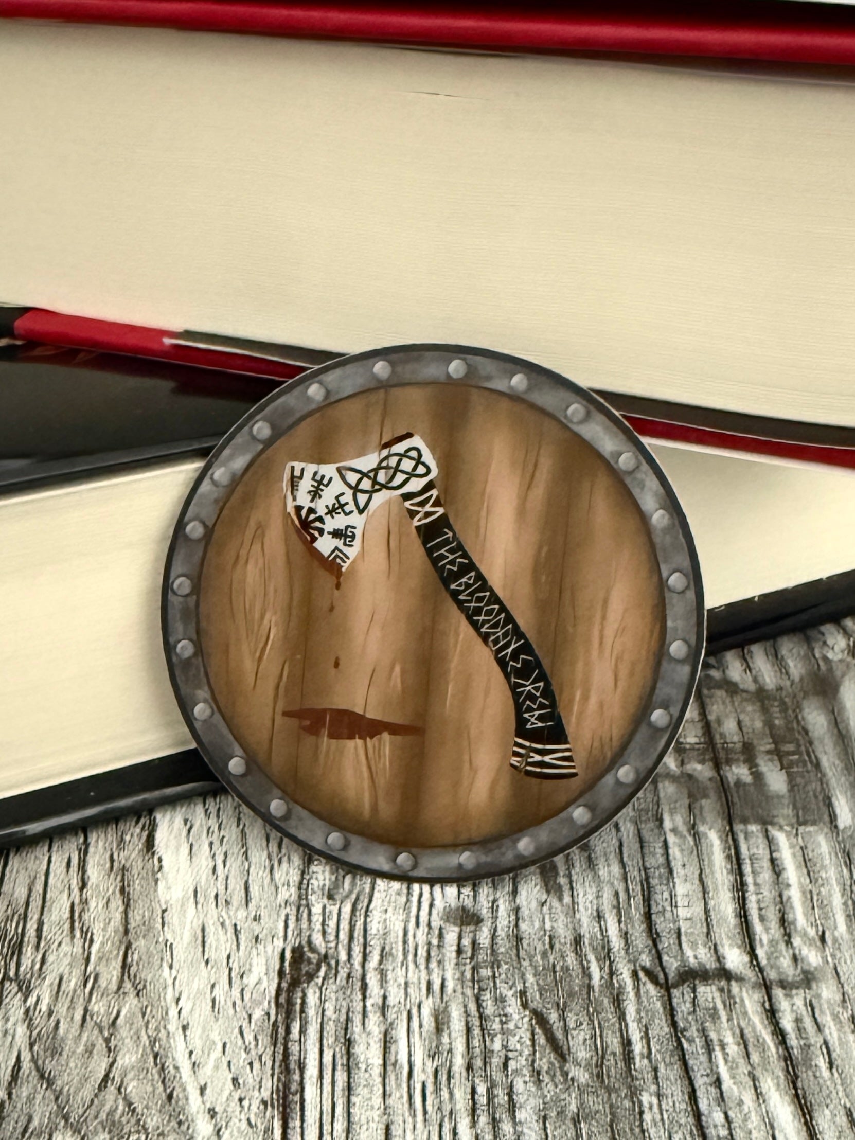 Round viking shield die cut sticker featuring an axe that reads The Bloodaxe Crew in runes down the handle, leaning against books, inspired by The Road of Bones by Demi Winters