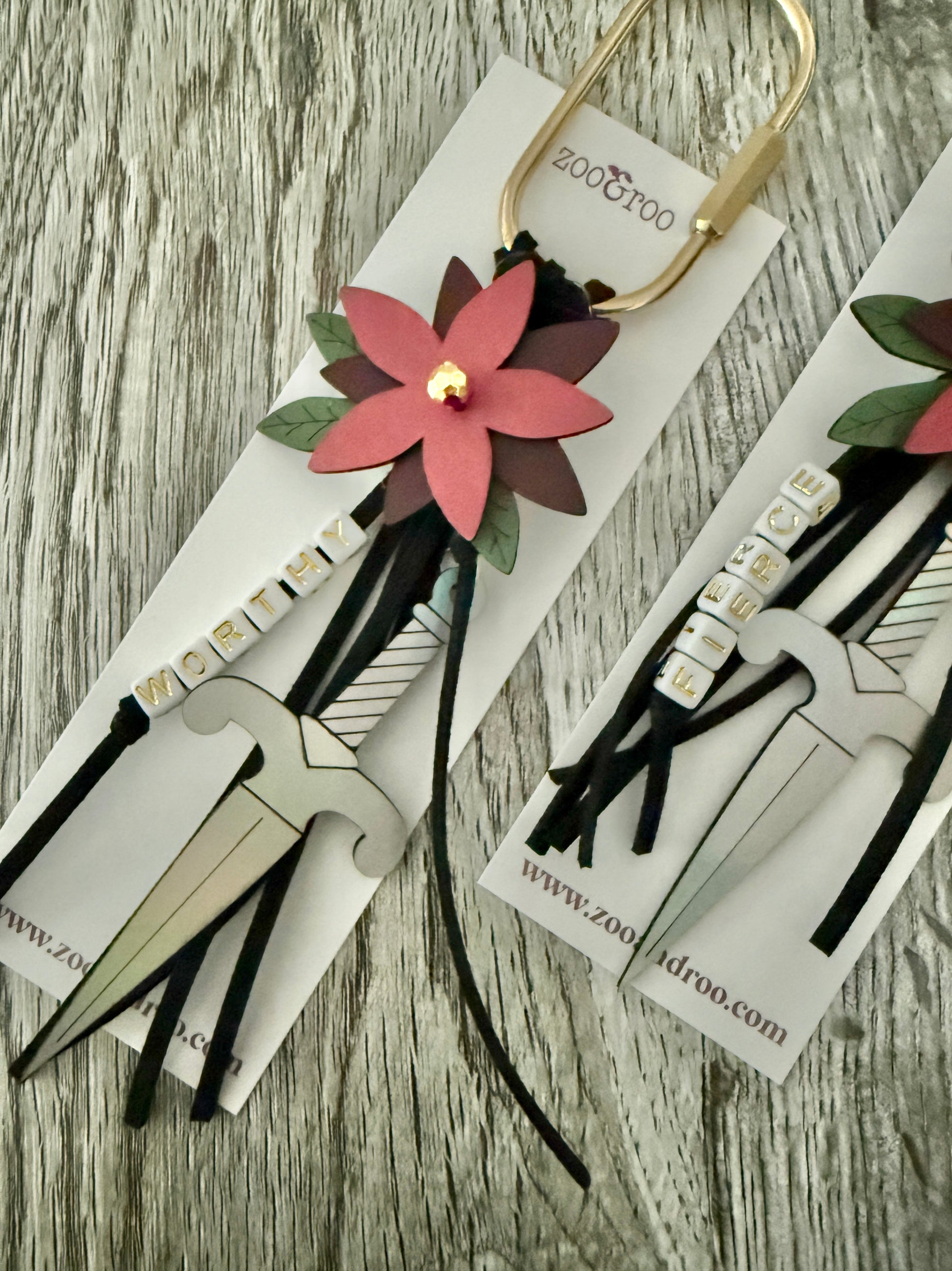 Bloom & Dagger romantasy bag charms by zoo&roo with layered burgundy vegan leather flower, iridescent dagger charm, green leatherette leaves, and words WORTHY and FIERCE spelled out in beads on gold brass carabiner