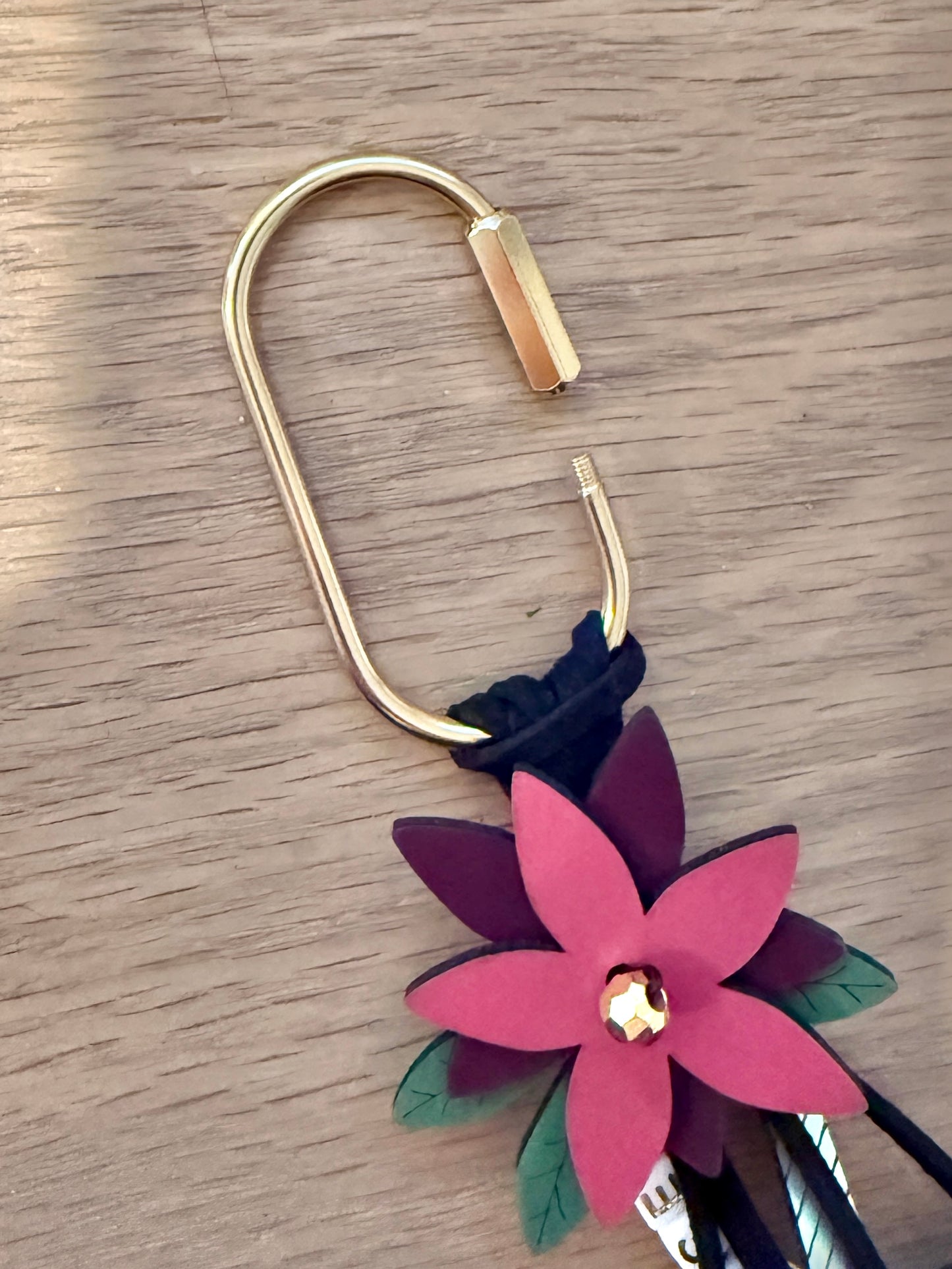 Gold caribiner with black cording and a pink and purple leather flower hanging from it as a bag charm