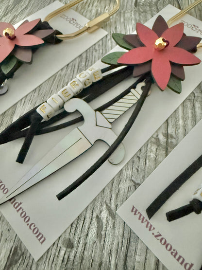Bloom & Dagger romantasy bag charm by zoo&roo with black suede cords, layered burgundy vegan leather flower, iridescent dagger charm, green leatherette leaves, and FIERCE spelled out in beads on gold brass carabiner